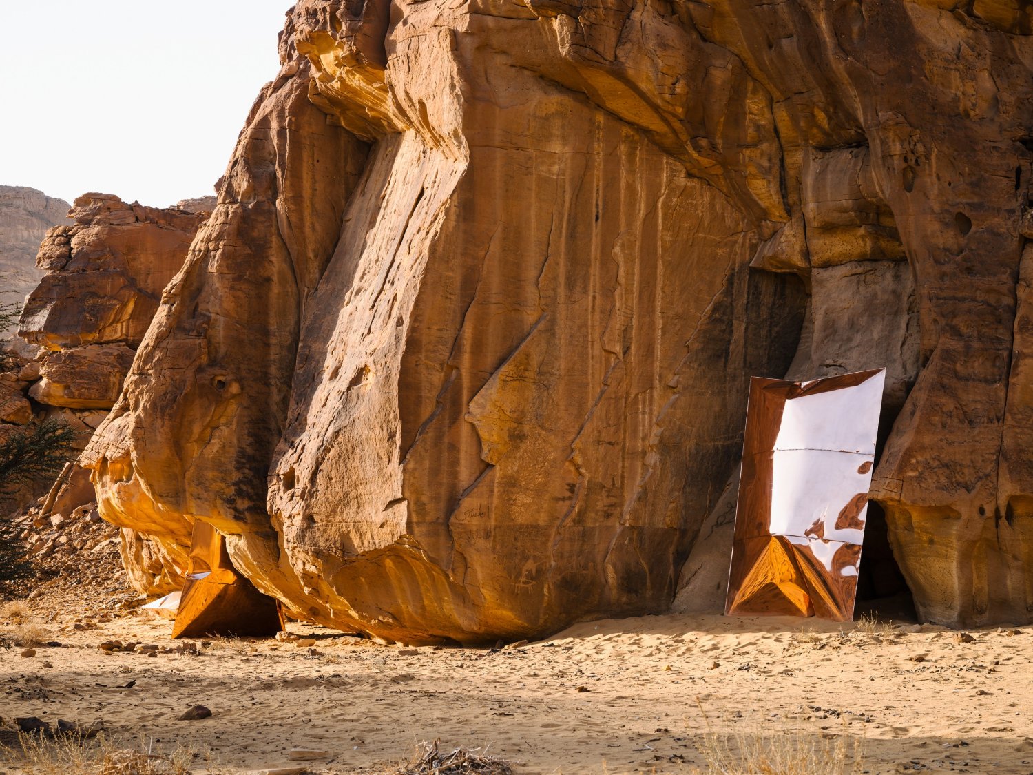 Desert X AlUla 2022 Group Exhibition — A monumental sculptural installation anchors the desert floor against a naturally stratified sandstone cliff face.