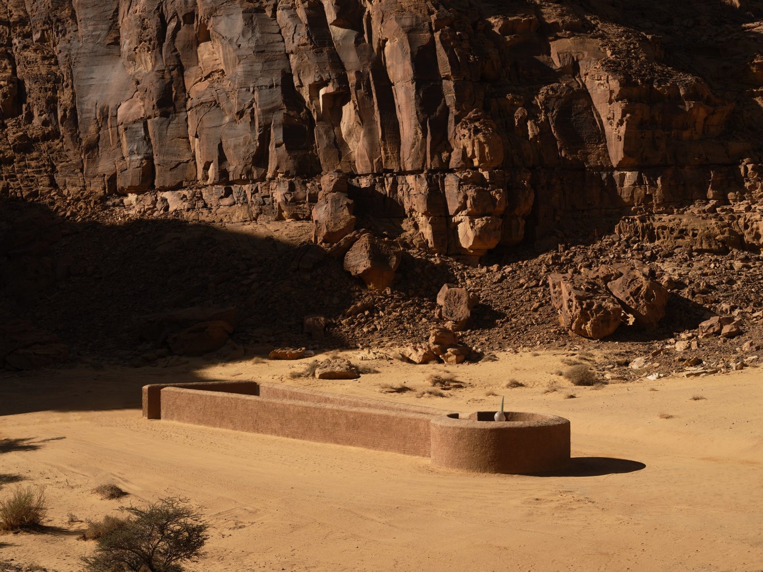 Desert X AlUla 2022 Group Exhibition — A monolithic cast-concrete bench with densely patterned surface reliefs sits on the sandy desert floor, positioned against a dramatically eroded sandstone...