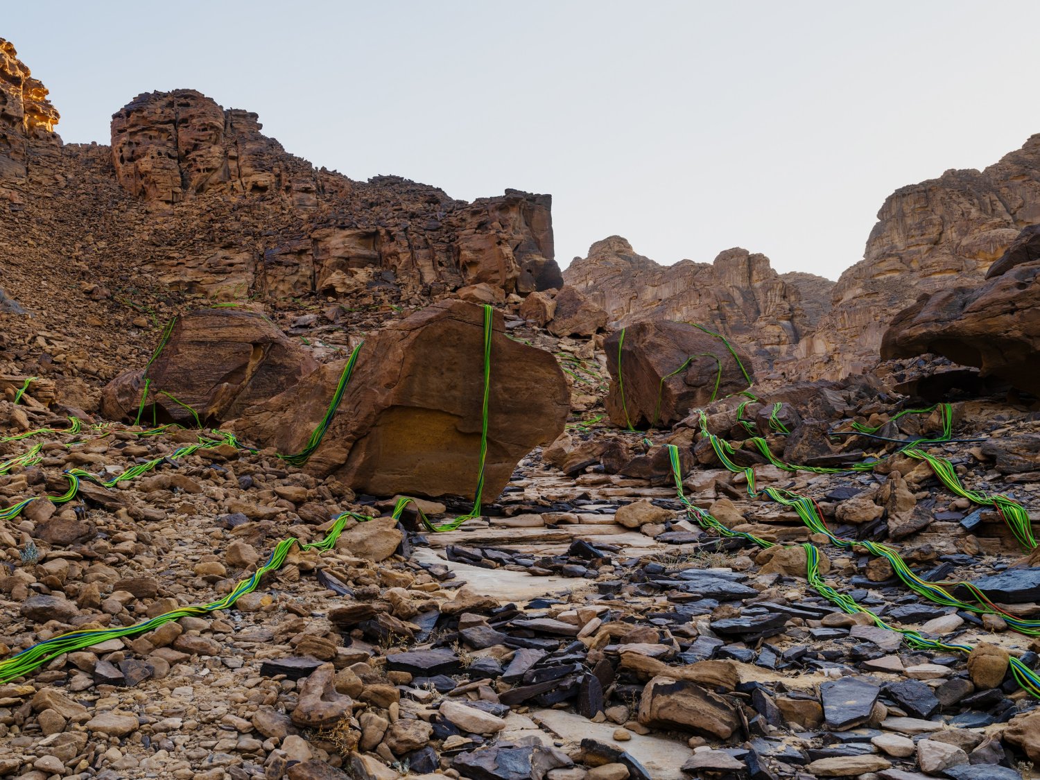 Desert X AlUla 2022 Group Exhibition — A site-specific installation of bright green, linear forms spiraling across boulders and rocky terrain in a desert wadi.