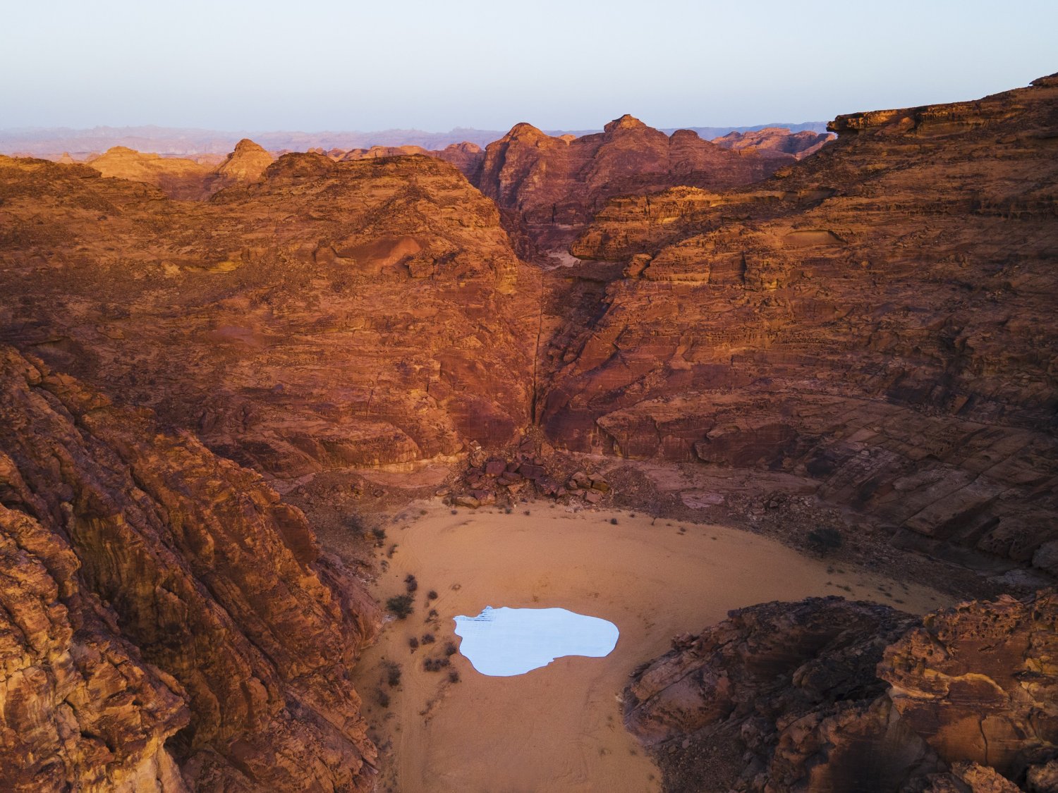 Desert X AlUla 2022 Group Exhibition — A sunlit pool of water reflects the pale desert sky, creating an optical inversion within the sandstone canyon basin.