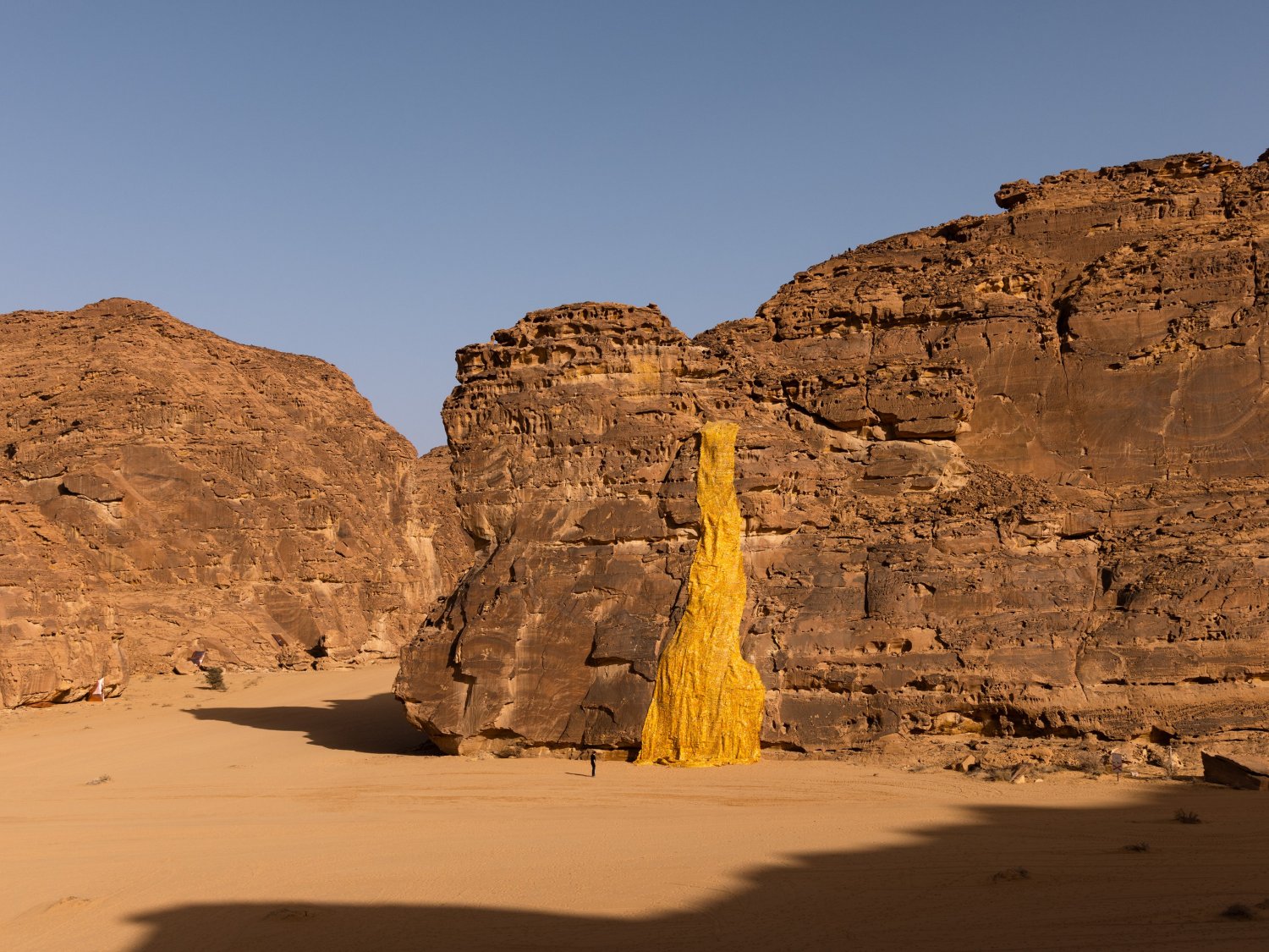 Desert X AlUla 2022 Group Exhibition — A monumental cast-brass or bronze sculptural form dominates the desert floor, its surface densely covered with repeating spiral ammonite reliefs.
