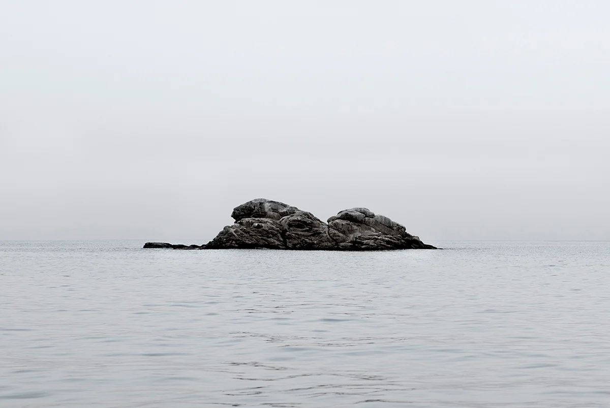 Island Hoping by Christina Dimitriadis — A solitary granite rock formation rises from calm Mediterranean waters, its surface marked by pronounced weathering and fracture patterns.