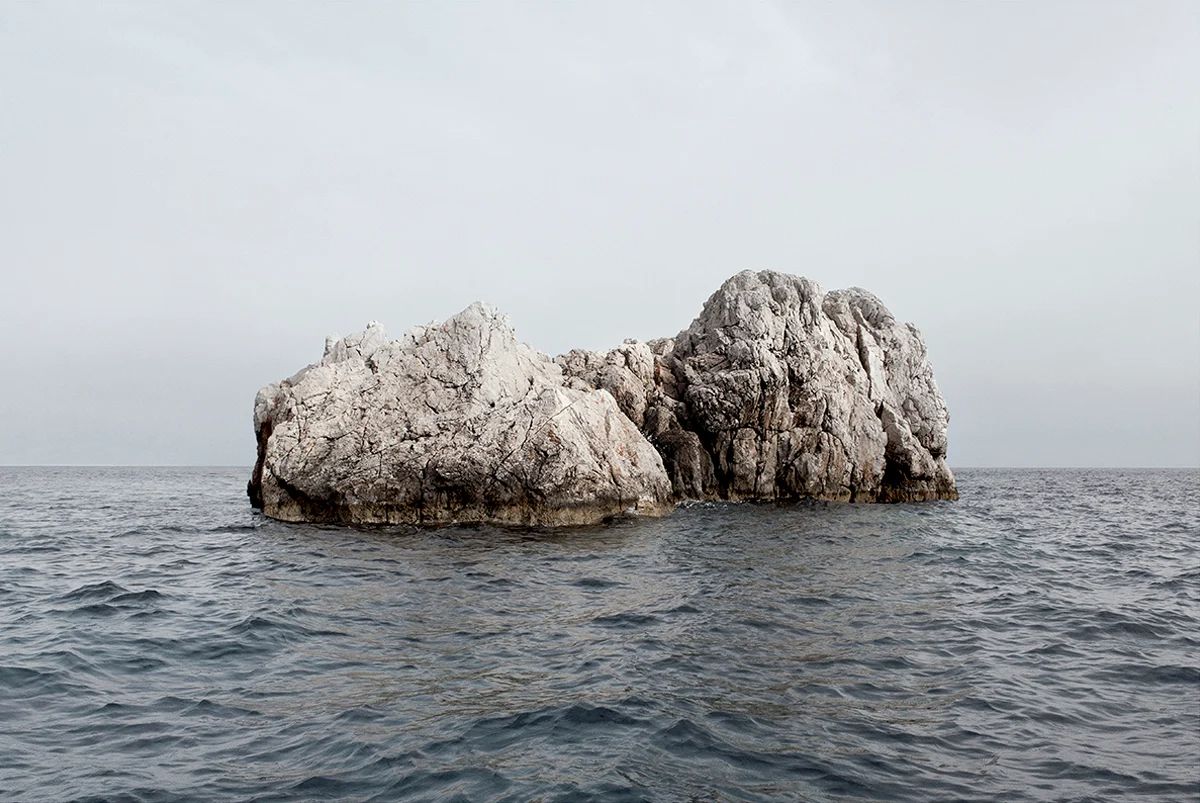Island Hoping by Christina Dimitriadis — A limestone or chalk formation rises from calm Mediterranean waters, its surface weathered into pronounced vertical striations and columnar breaks.
