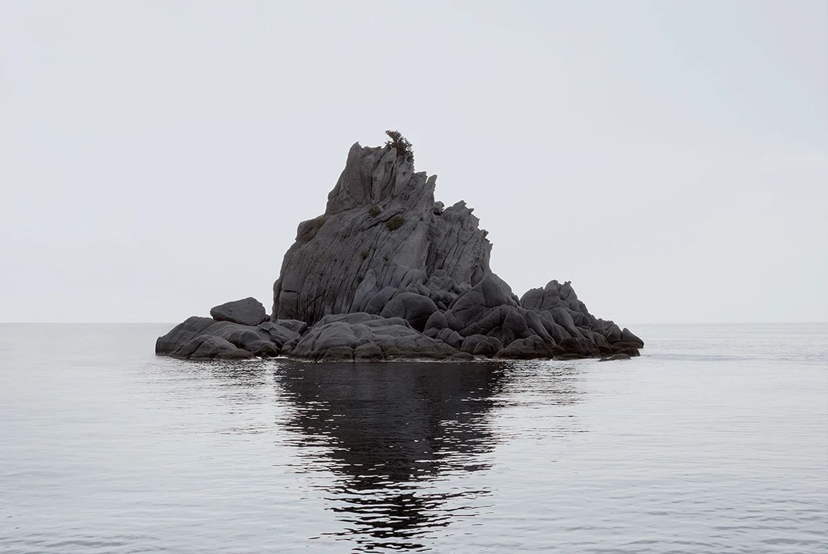 Island Hoping by Christina Dimitriadis — A columnar basalt formation rises from calm Mediterranean waters, its surface defined by pronounced vertical striations that create a rhythmic pattern...