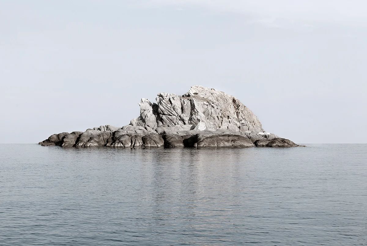Island Hoping by Christina Dimitriadis — A solitary granite rock formation rises from calm Mediterranean waters, its upper structure defined by pronounced columnar basalt striations and jagged...