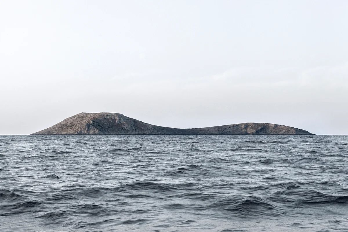 Island Hoping by Christina Dimitriadis — A single rocky island formation rises from grey-blue water, its elongated profile characterized by columnar basalt structures with visible vertical...