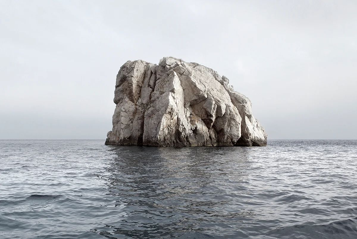 Island Hoping by Christina Dimitriadis — A monolithic limestone formation rises from calm Mediterranean waters, its weathered surface defined by deep vertical striations and pronounced columnar...