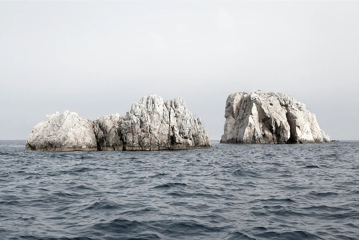 Island Hoping by Christina Dimitriadis — Two limestone rock formations rise distinctly from calm Mediterranean waters, their pale stone surfaces marked by deep vertical striations and weathered...