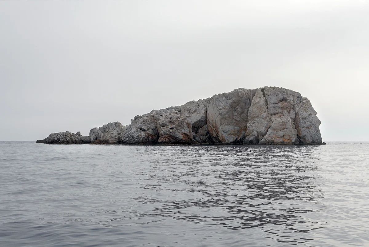 Island Hoping by Christina Dimitriadis — A limestone or chalk rock formation rises from calm Mediterranean waters, its surface marked by deep vertical fractures and columnar striations.