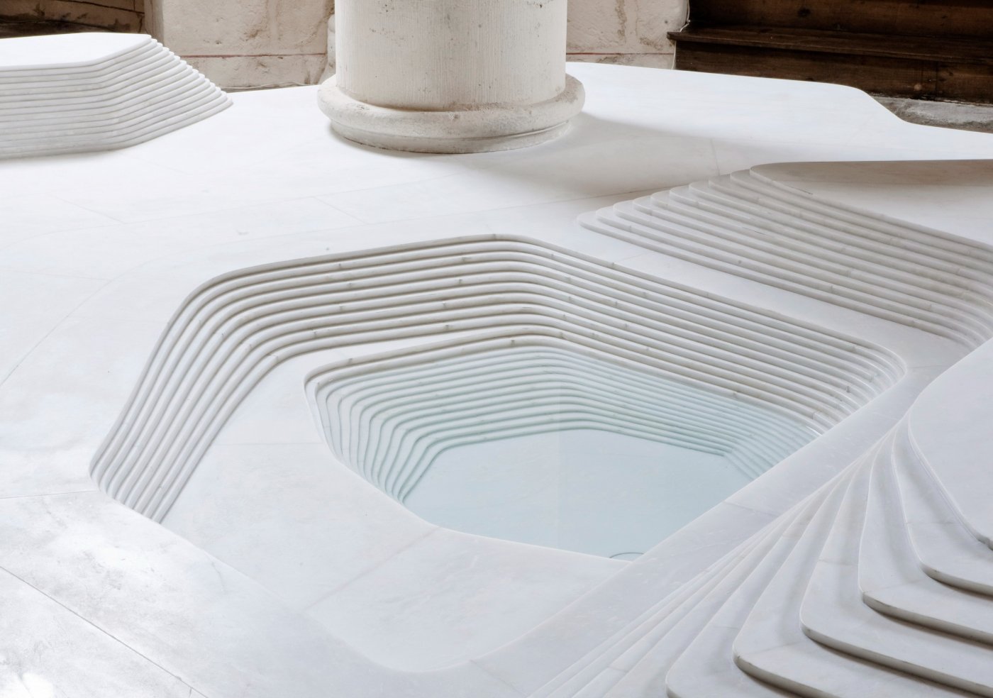 St Hilaire Church in Melle by Mathieu Lehanneur — Close-up detail of the baptistery basin carved from white marble, revealing the layered construction technique through concentric rippled patterns that...