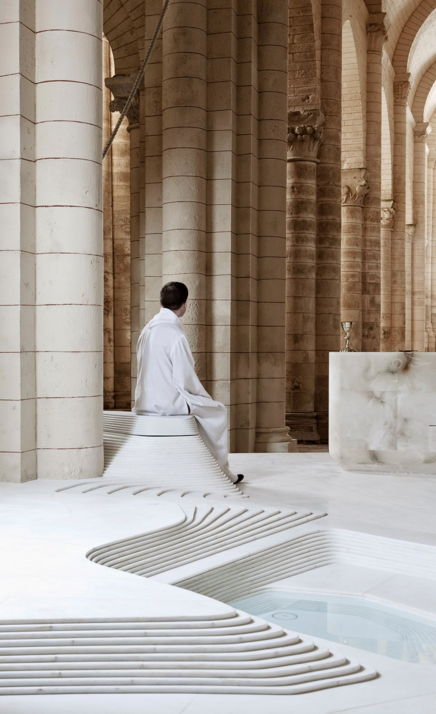 St Hilaire Church in Melle by Mathieu Lehanneur — A figure in white linen moves through the Romanesque interior, passing between cylindrical stone columns with carved capitals.