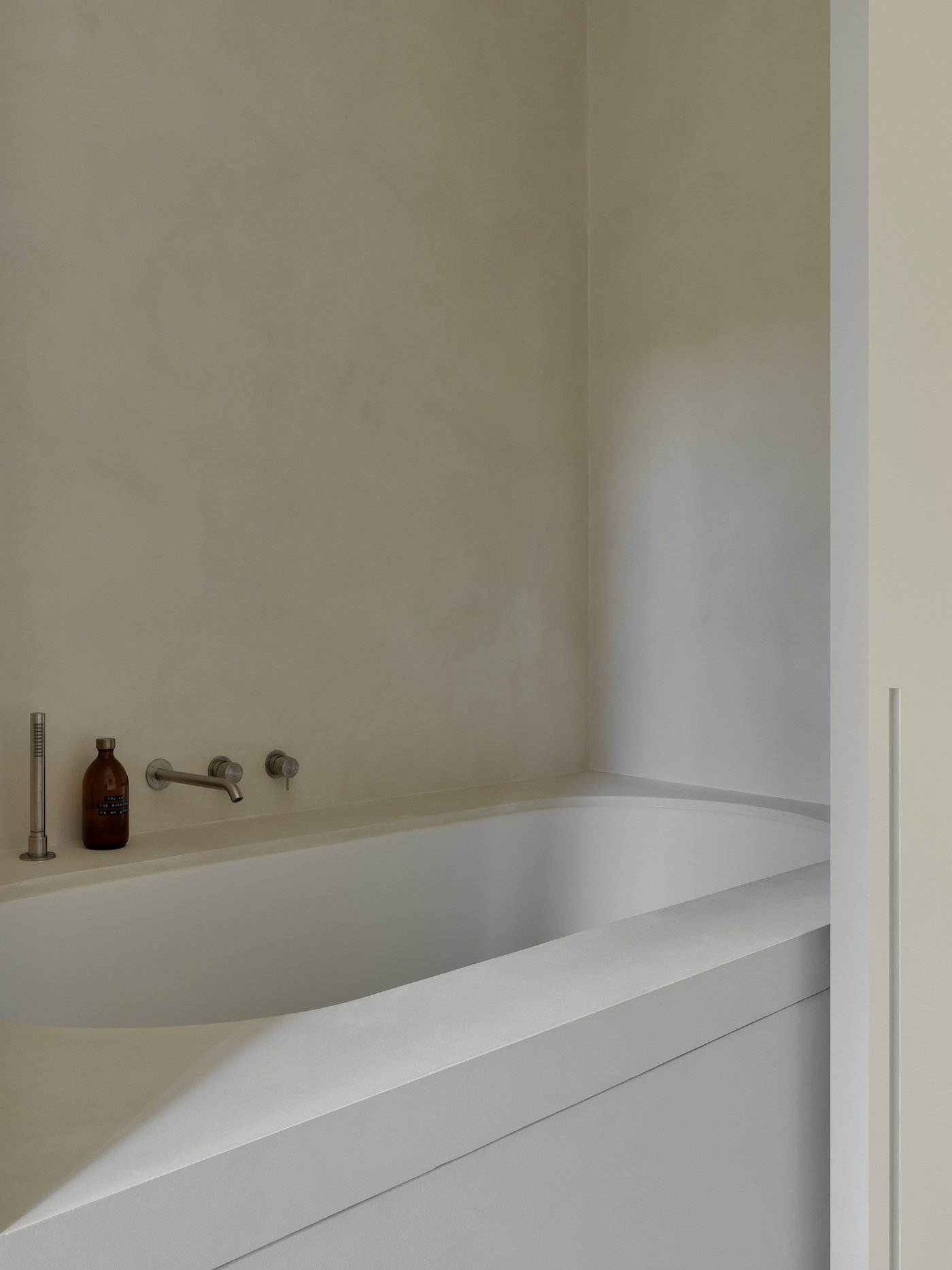Villa BA by gosseye + verbeke — A minimalist bathroom alcove carved into white plaster walls, featuring a sculptural sunken bathtub with curved edges and a continuous rim.