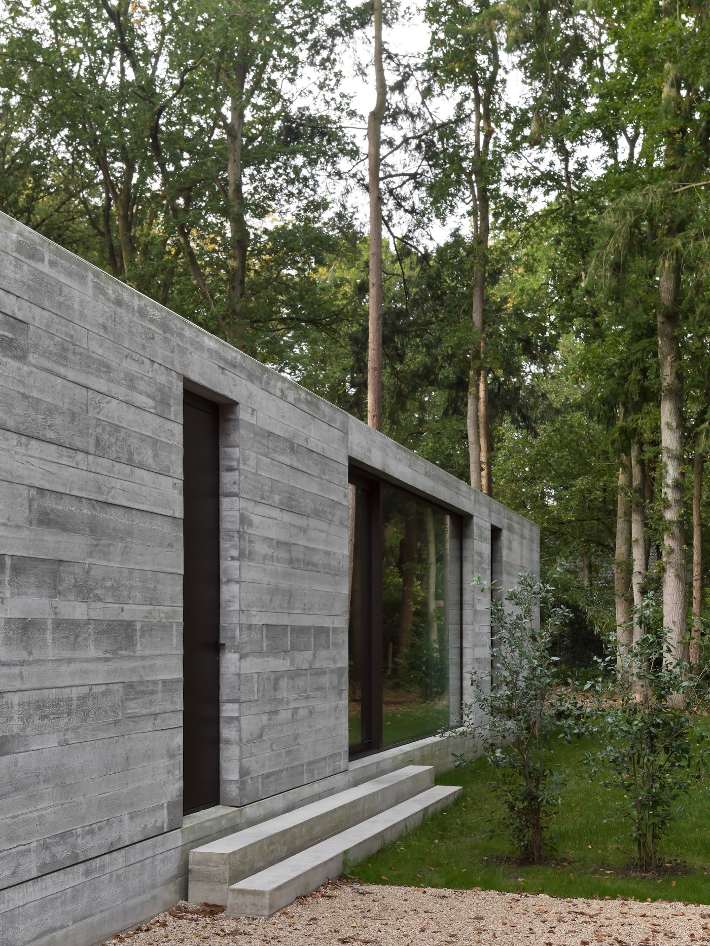 Villa BA by gosseye + verbeke — Villa BA's entrance facade presents a board-formed concrete wall punctuated by a centered black door and large glazed opening, creating a studied symmetry.