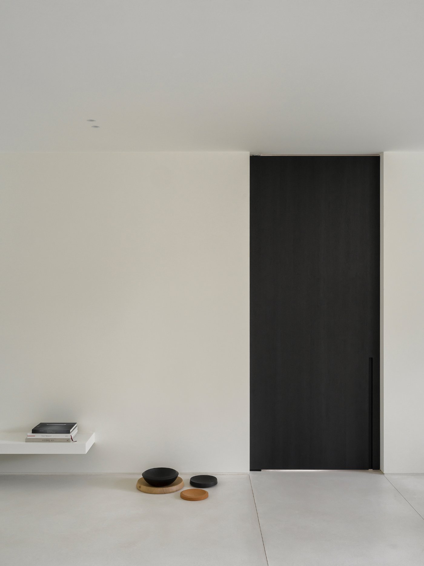 Villa BA by gosseye + verbeke — A centered black door with vertical handle punctuates the concrete facade, framed by symmetrical grey planes.