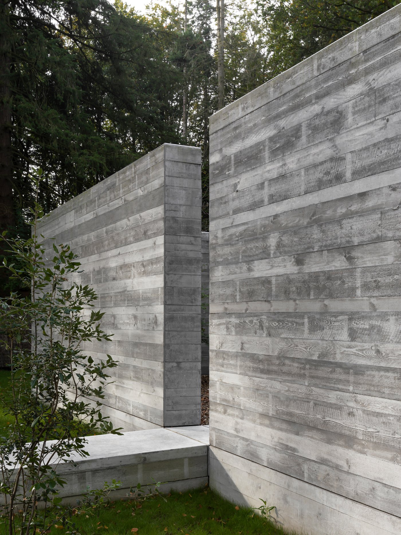 Villa BA by gosseye + verbeke — A concrete passage frames the entry to Villa BA, its board-formed walls displaying horizontal wood-grain textures in grey and white striations.