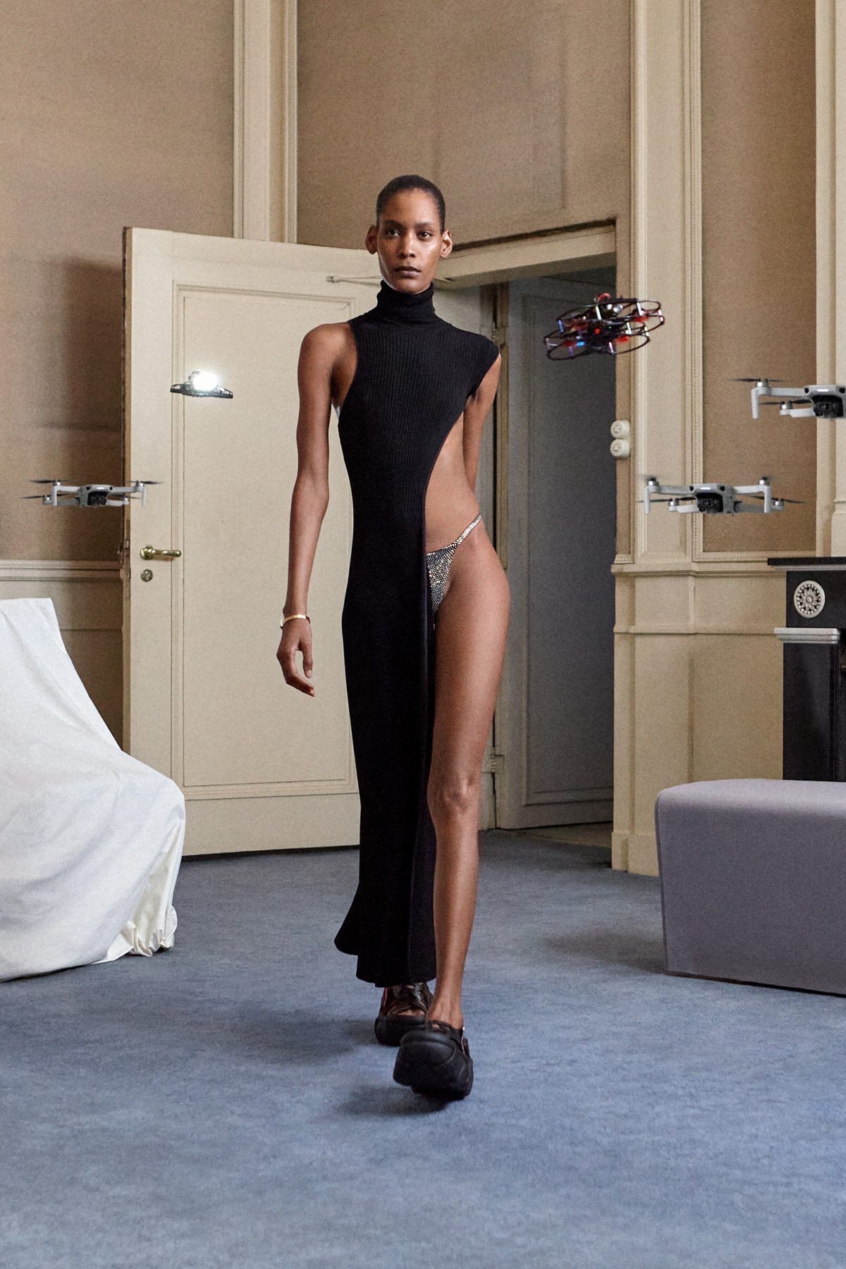 SSAW22 by Duran Lantink — A model wears a black ribbed-knit sleeveless turtleneck dress with dramatic asymmetrical cutouts exposing the torso and right leg, paired with a crystalline...