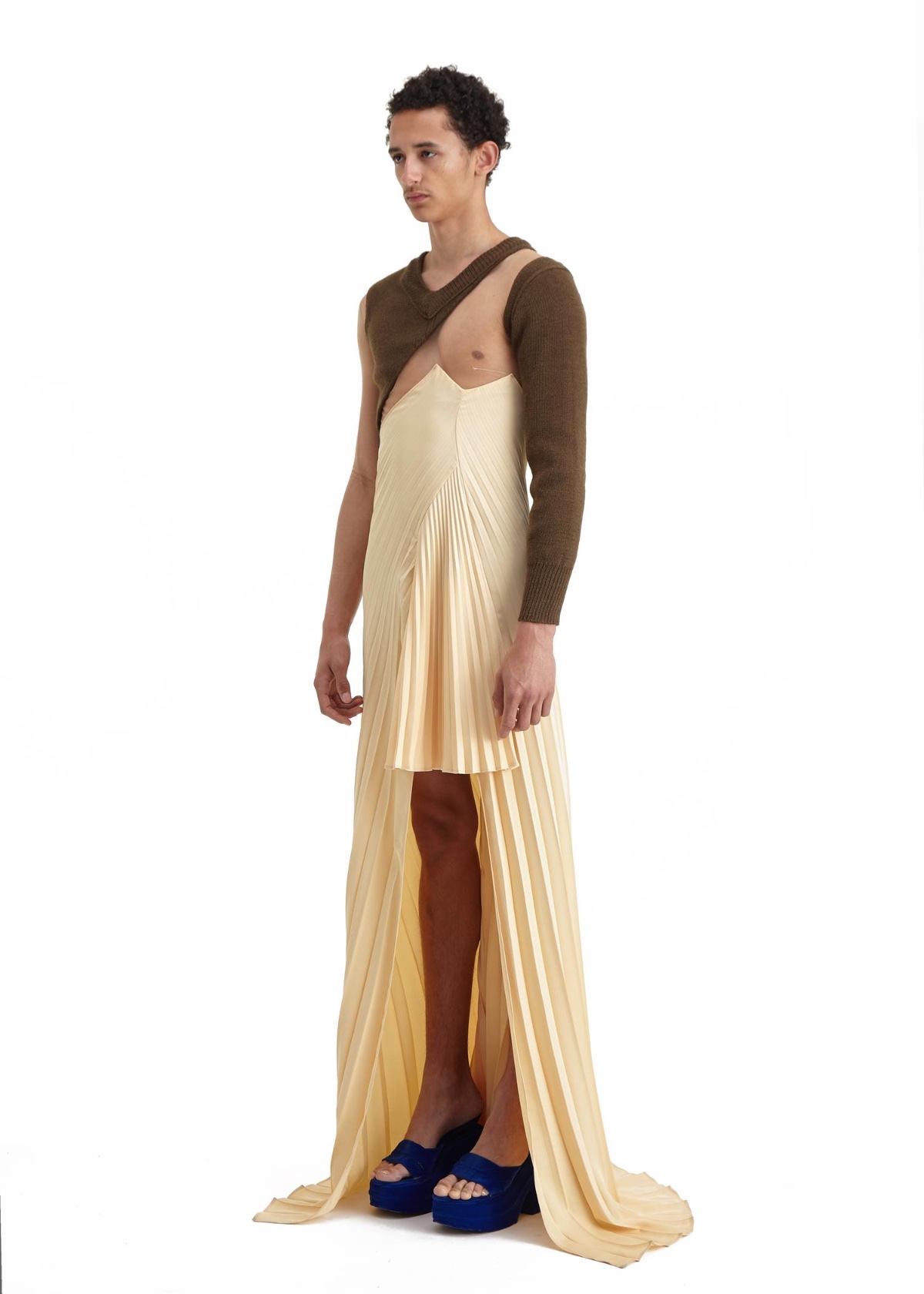 SSAW22 by Duran Lantink — A model wearing a cream pleated floor-length dress with asymmetrical cutouts reveals one leg and the torso beneath a brown knit sleeve.