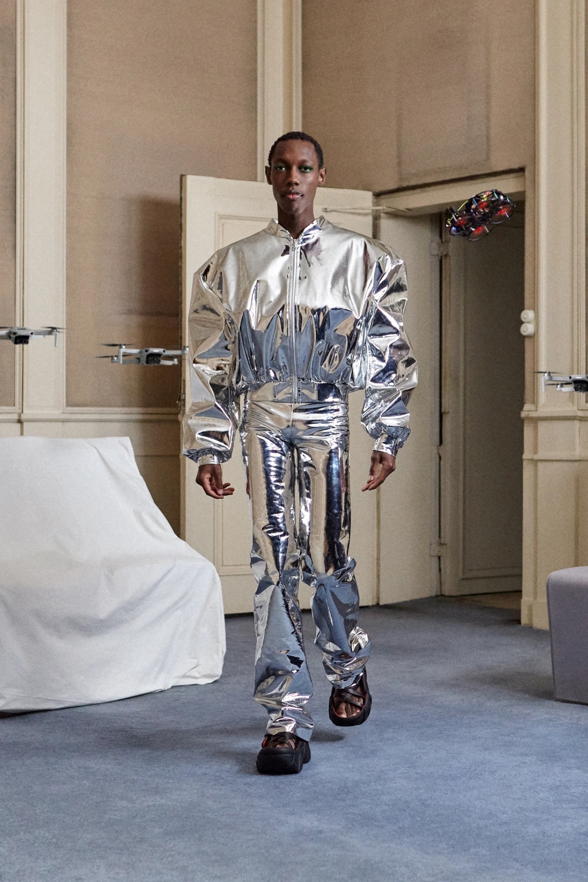 SSAW22 by Duran Lantink — A model wears a silver metallic two-piece ensemble composed of a reflective jacket and matching pants, both constructed from iridescent material with...