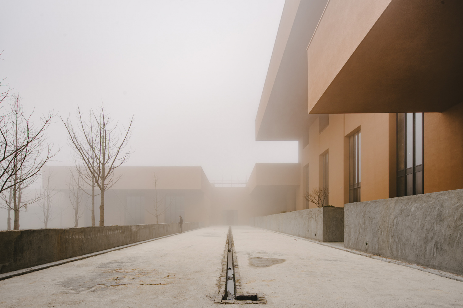 Zhejiang Museum of Natural History by David Chipperfield Architects — A symmetrical portal framed by two parallel rendered concrete walls in warm terracotta tone extends toward a forested hillside shrouded in mist.