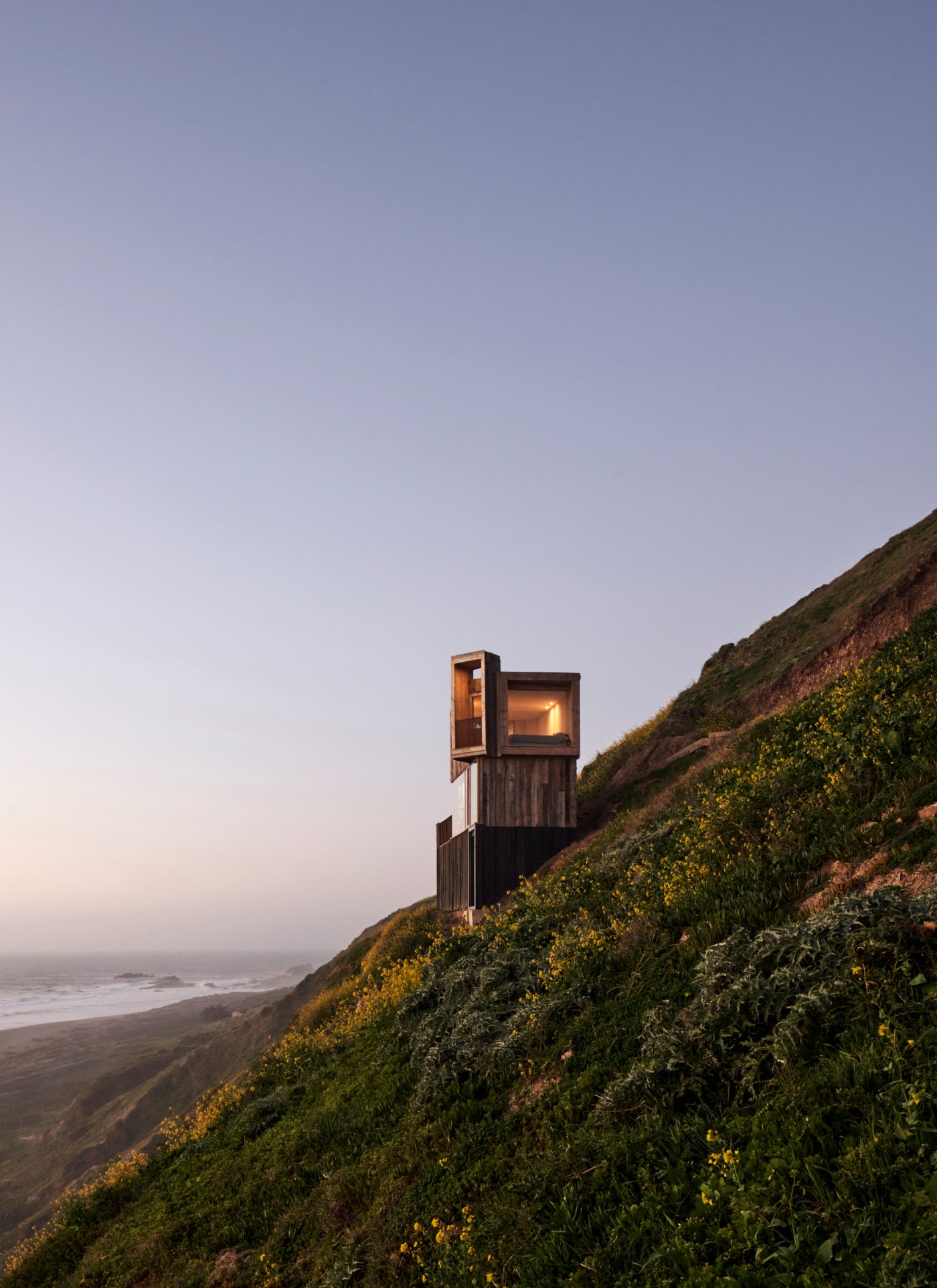 La Loica and La Tagua Cabins by Croxatto y Opazo Arquitectos by Croxatto and Opazo Architects — A reclaimed oak cabin rises from a coastal hillside densely planted with yellow wildflowers and scrub vegetation, its weathered dark timber cladding visible...