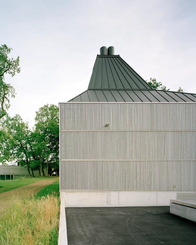 District Heating Plant by MAK Architecture — The District Heating Plant presents its volumetric complexity through a low-angle view that emphasizes the dual cylindrical chimneys rising from a pyramidal...