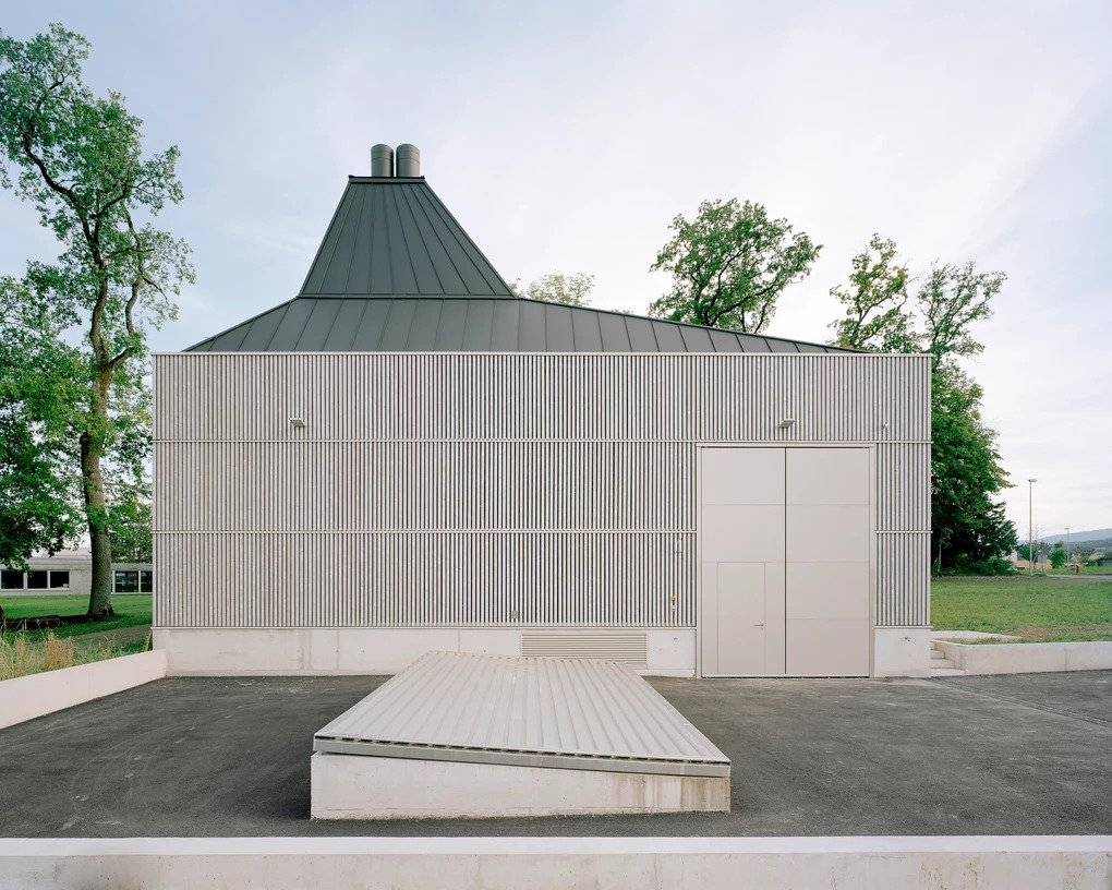 District Heating Plant by MAK Architecture — The District Heating Plant presents a geometric composition centered on a pyramidal roof form crowned with dual cylindrical chimneys.