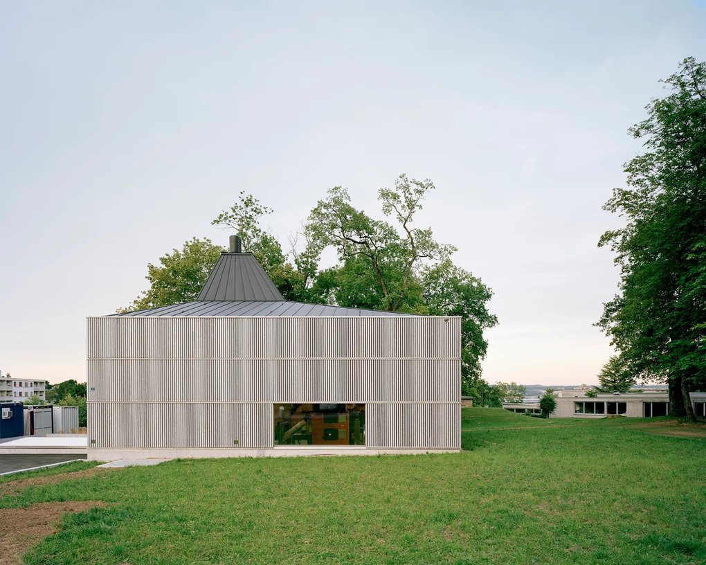 District Heating Plant by MAK Architecture — A corrugated metal heating plant sits within residential greenery, its boxy rectilinear volume anchored by a concrete plinth.