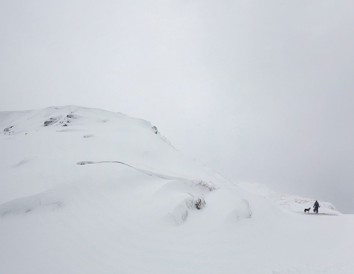 The Weather Report by Marzena Skubatz — A solitary figure with a dog traverses a snow-covered ridge in Iceland's remote Marsibil region.
