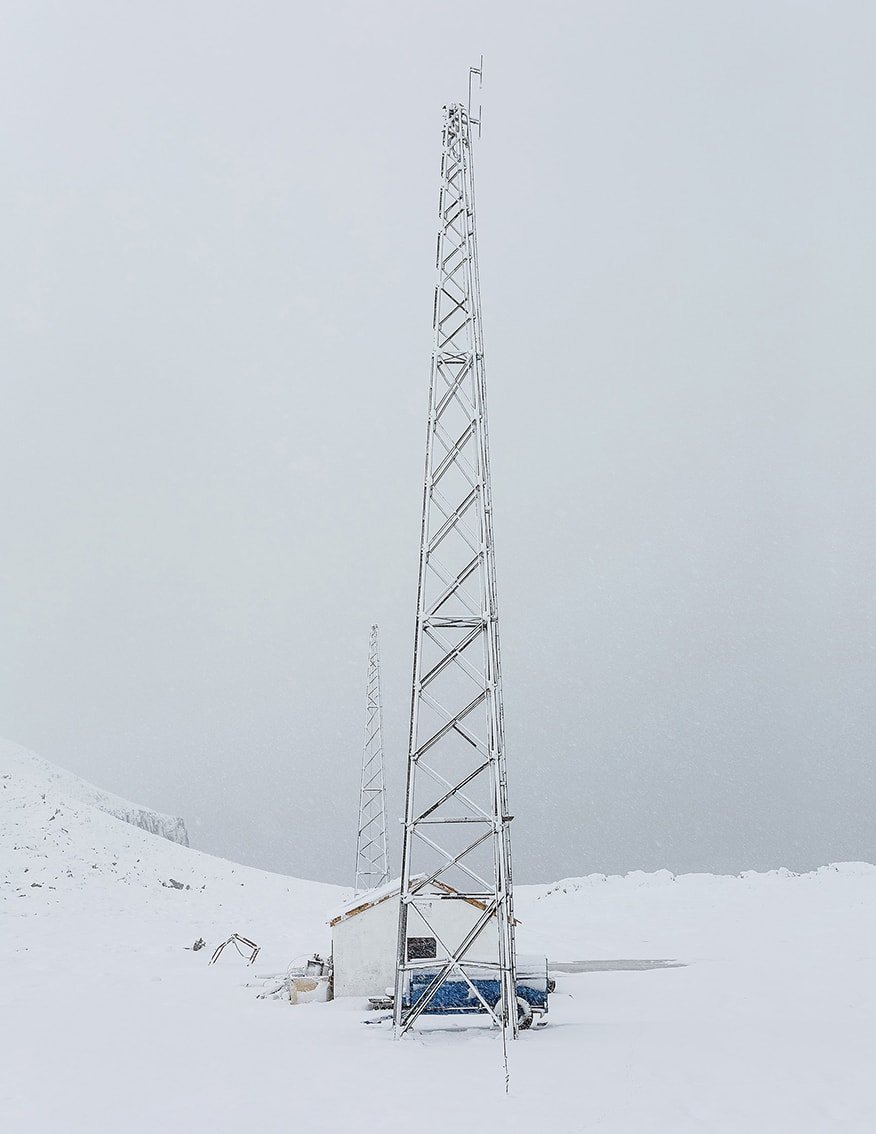 The Weather Report by Marzena Skubatz — A tall lattice communications tower dominates the snow-covered landscape of Marsibil, Iceland, its geometric steel framework rendered monochromatic against...