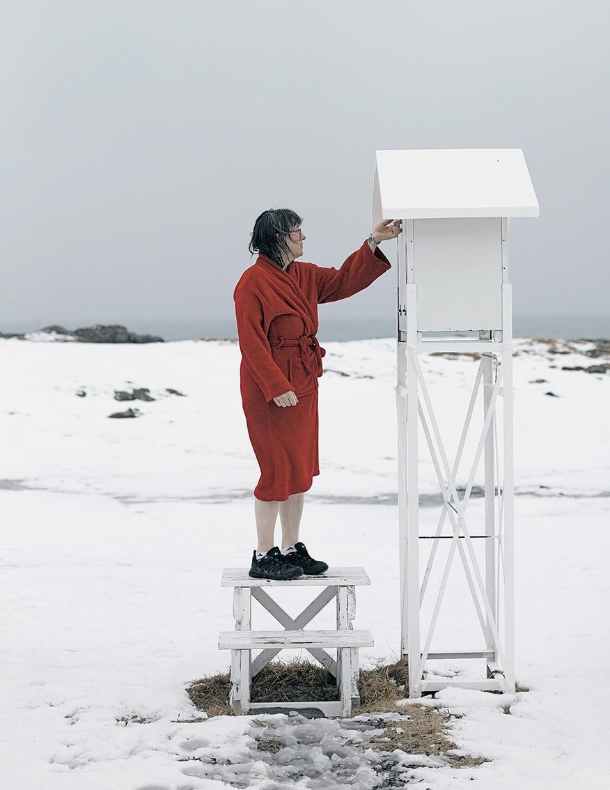 The Weather Report by Marzena Skubatz — A woman in a burgundy wool coat stands elevated on a timber-frame stepladder, tending to a white-painted metal weather station structure against an overcast...