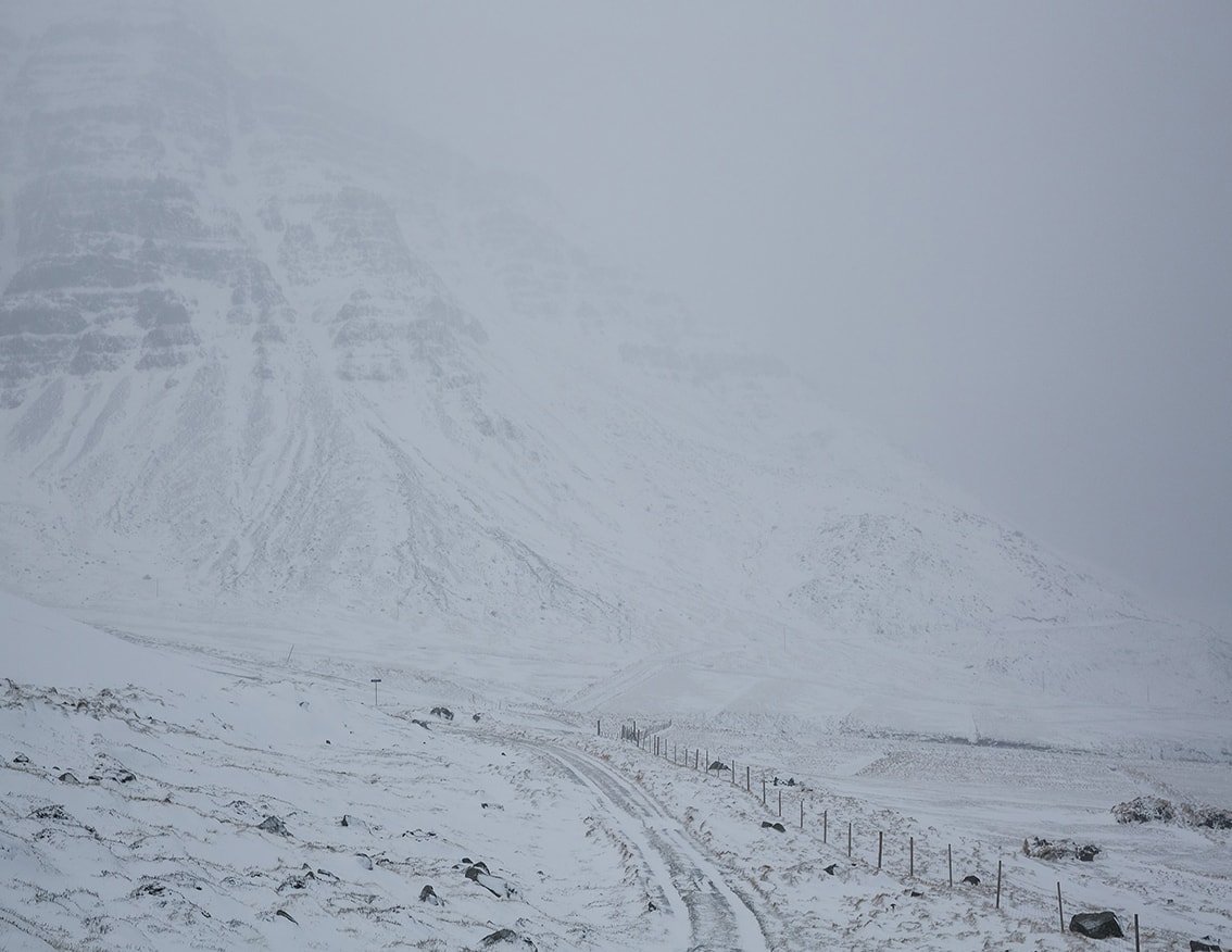 The Weather Report by Marzena Skubatz — A snow-covered Icelandic valley recedes into heavy precipitation and low visibility, with a single-track road marked by fence posts threading through the...