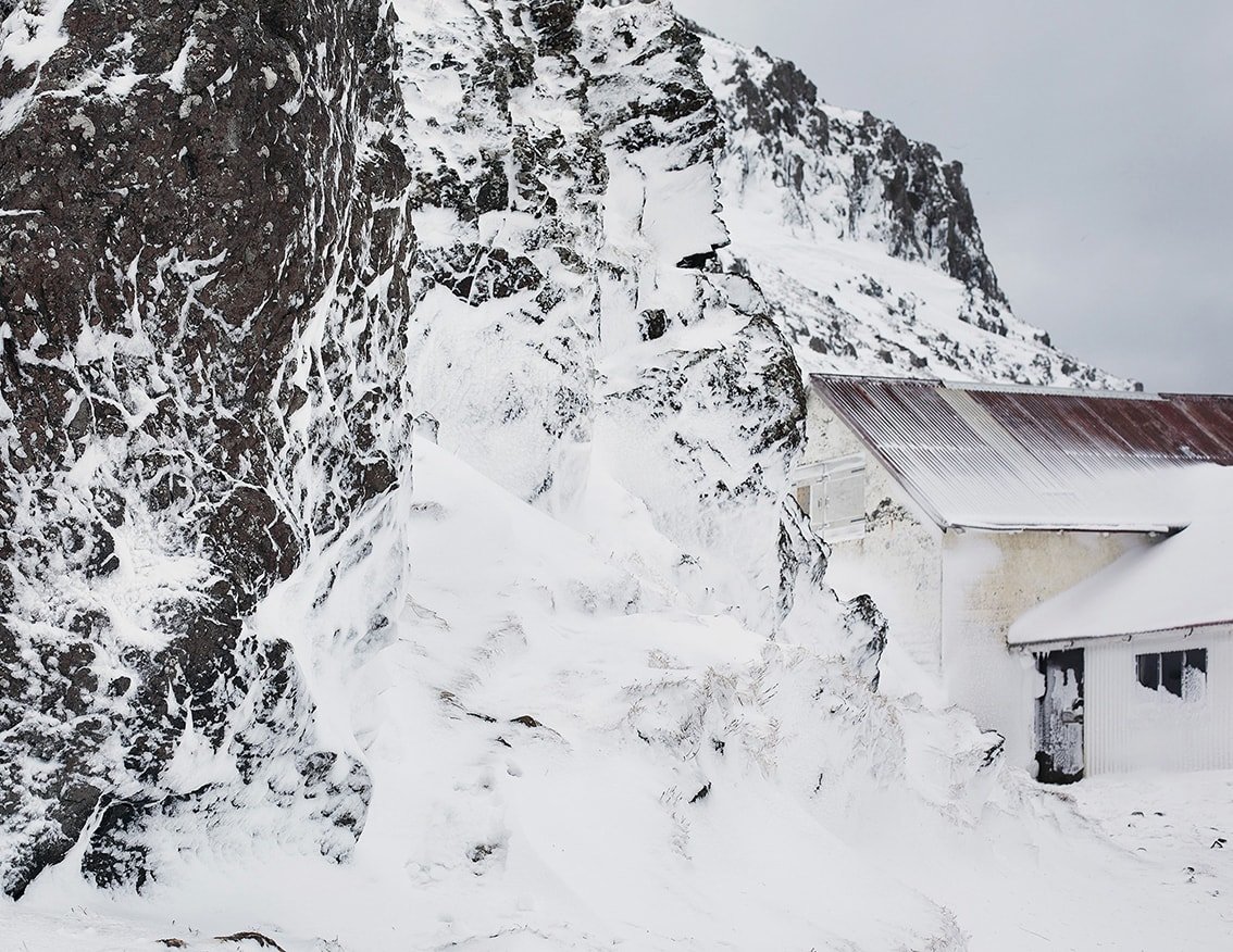 The Weather Report by Marzena Skubatz — A snow-buried timber-frame structure with corrugated metal roofing dominates the right frame, its rust-patinated panels contrasting against accumulated...