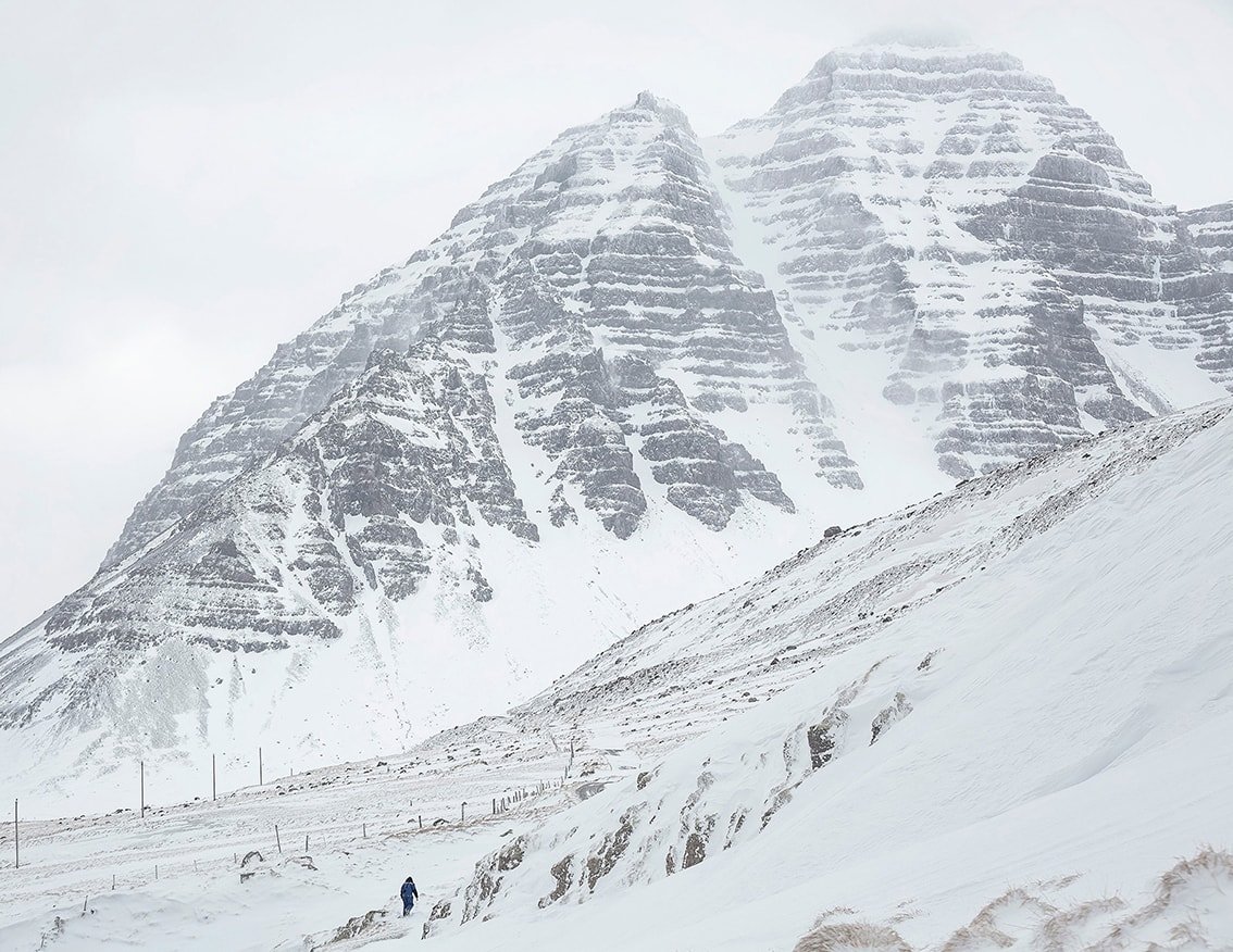 The Weather Report by Marzena Skubatz — A snow-covered mountainside dominates the composition, its stratified volcanic rock face rising steeply against an overcast sky.