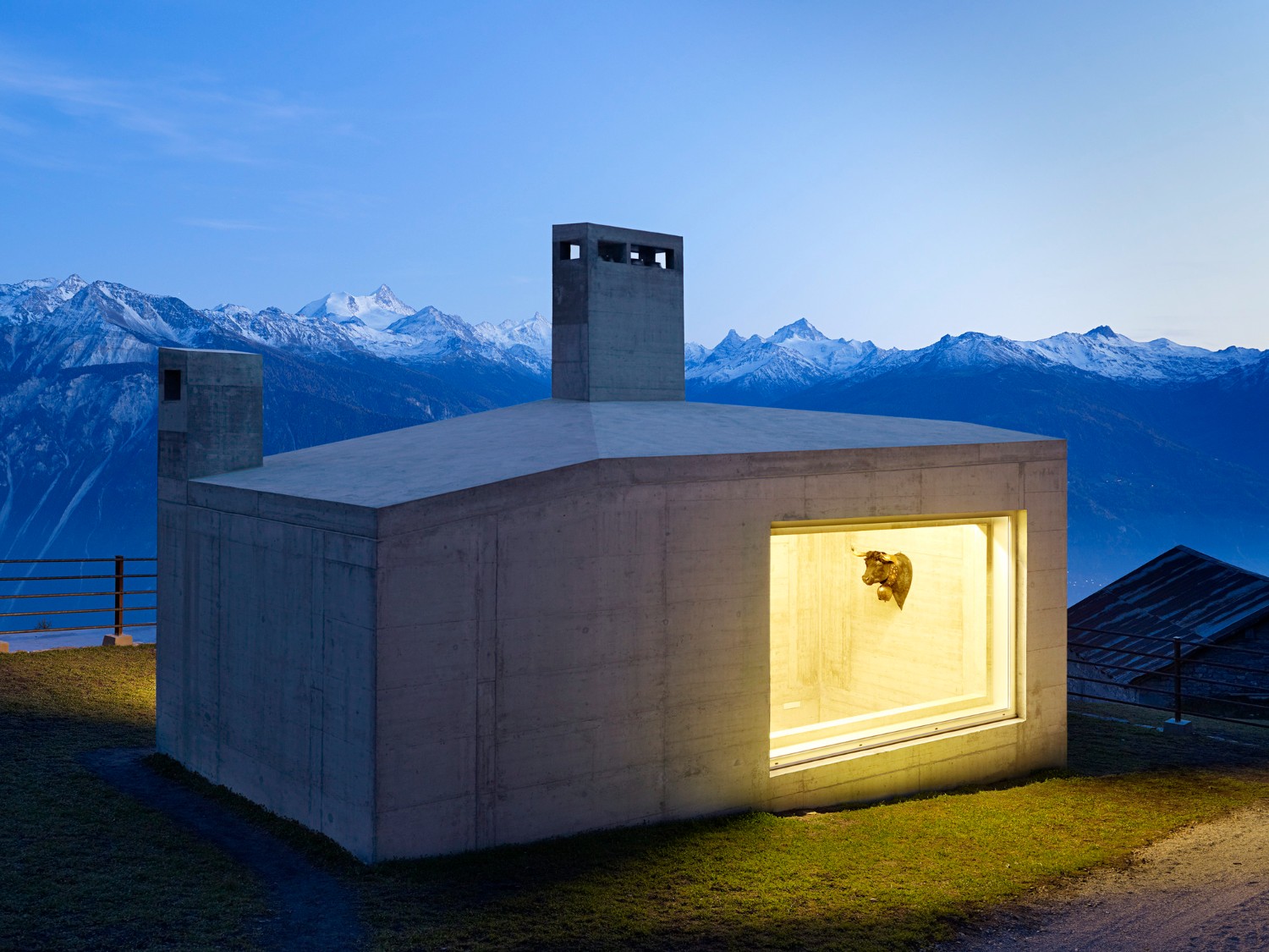 Community Shelter by frundgallina — A monolithic concrete shelter positioned on sloped alpine terrain, its mass divided into two distinct volumes at different elevations.