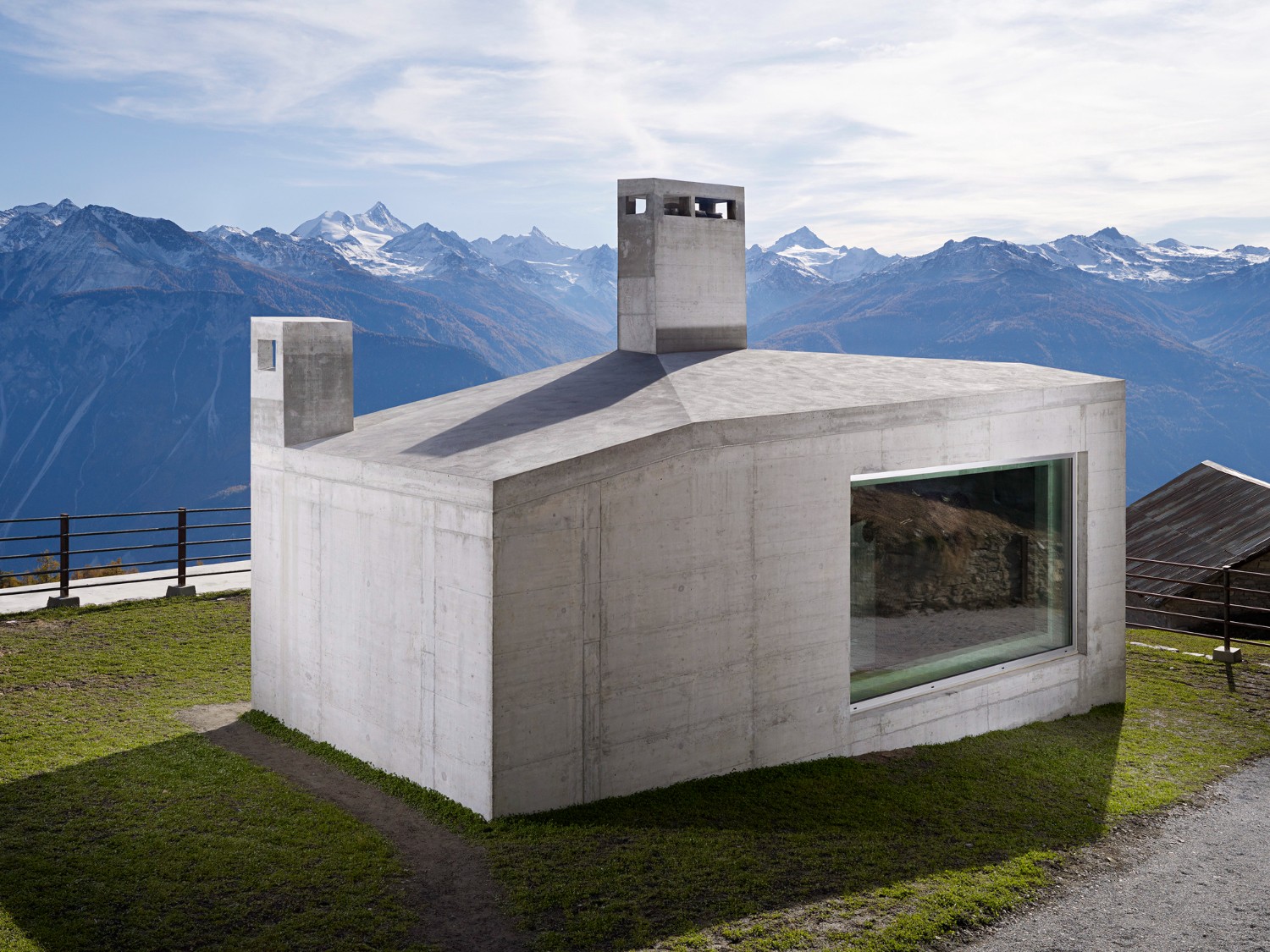 Community Shelter by frundgallina — A monolithic concrete shelter positioned on sloped alpine terrain, its cubic form divided by functional apertures.