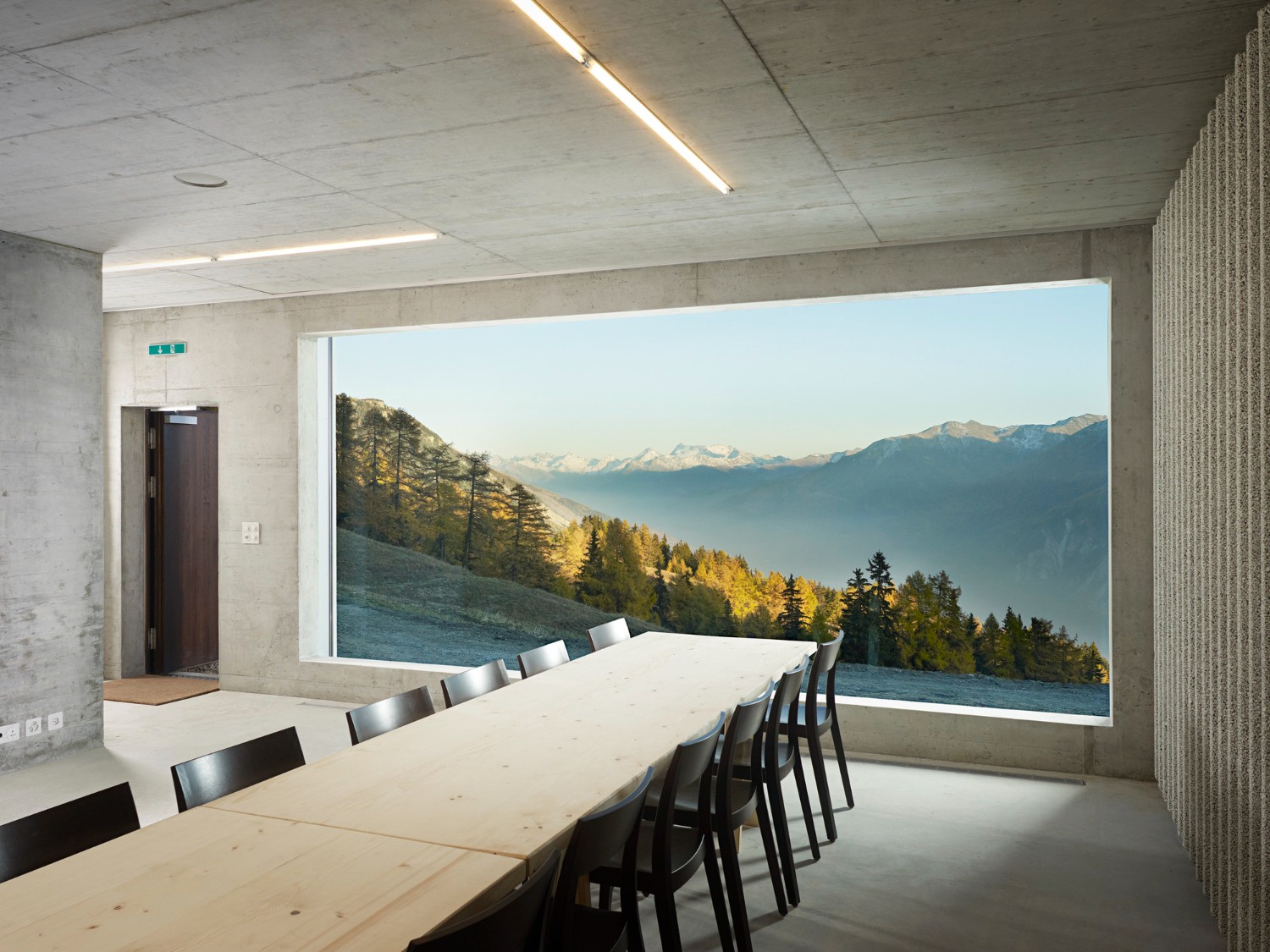 Community Shelter by frundgallina — A meeting room with board-formed concrete walls and ceiling houses a long timber table with black metal chairs aligned toward a monumental horizontal glazed...