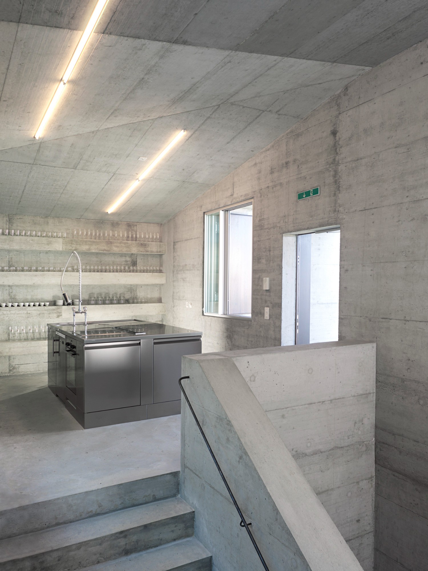 Community Shelter by frundgallina — Interior view of concrete shelter showing kitchen workspace with stainless steel sink and faucet positioned beneath shelving displaying glass vessels.