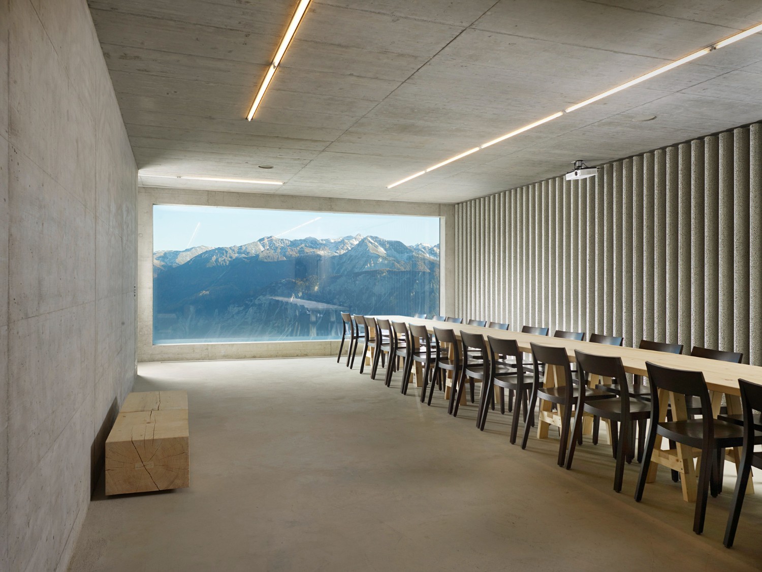 Community Shelter by frundgallina — A linear dining space defined by board-formed concrete ceiling and walls, flooded with natural light through a large horizontal glazed opening framing...