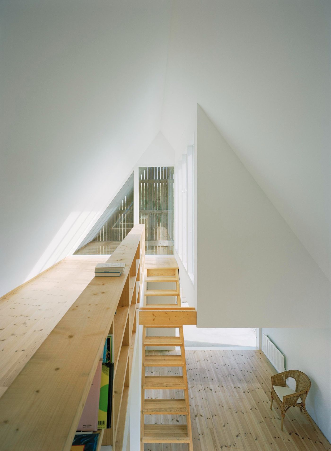 Sommarhus at Stora Gasmora by LLP Arkitektkontor — A wooden staircase with open pine treads ascends diagonally toward a skylit upper level, framed between white-painted angled walls.