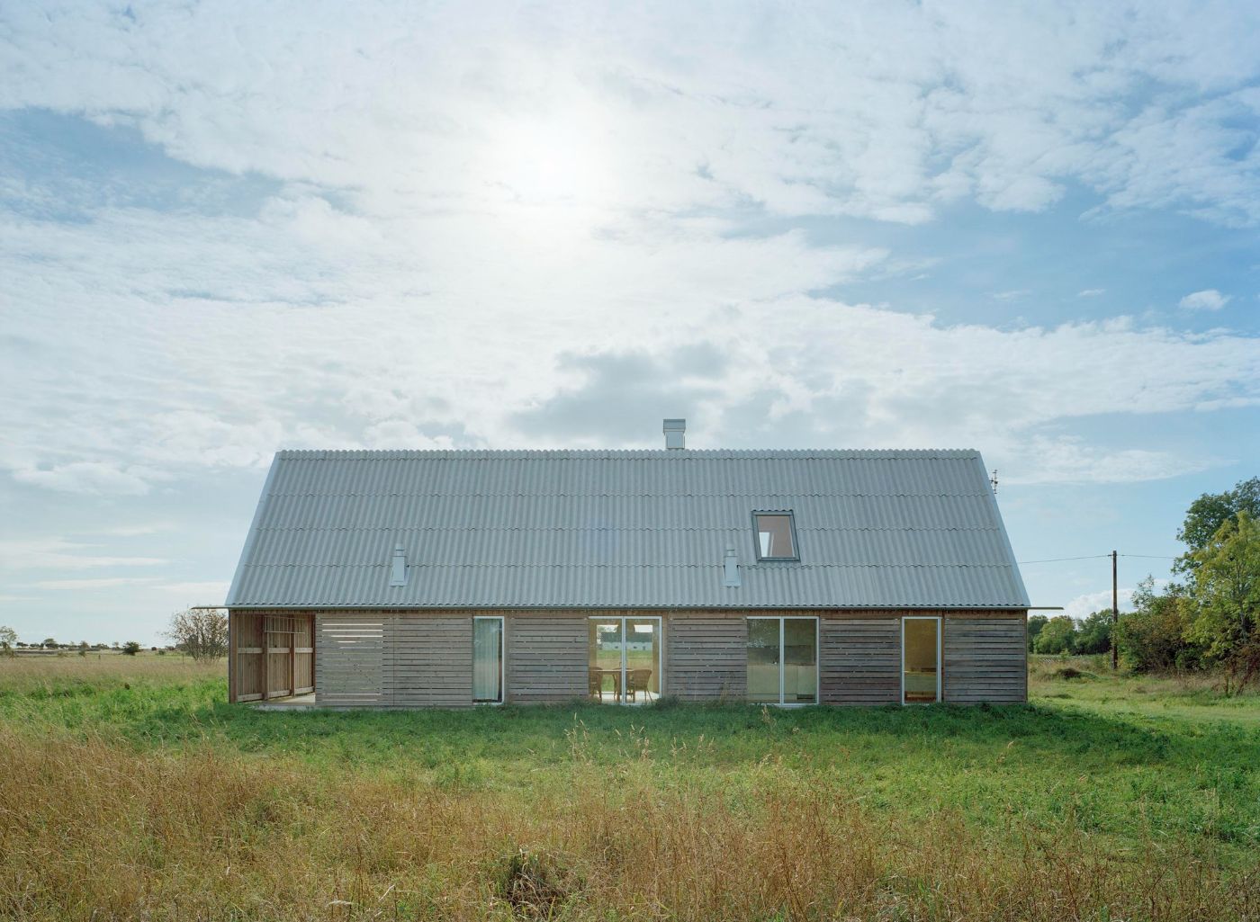 Sommarhus at Stora Gasmora by LLP Arkitektkontor — A single-story timber dwelling with a pitched fiber cement roof sits isolated within flat agricultural meadowland facing the Baltic Sea.