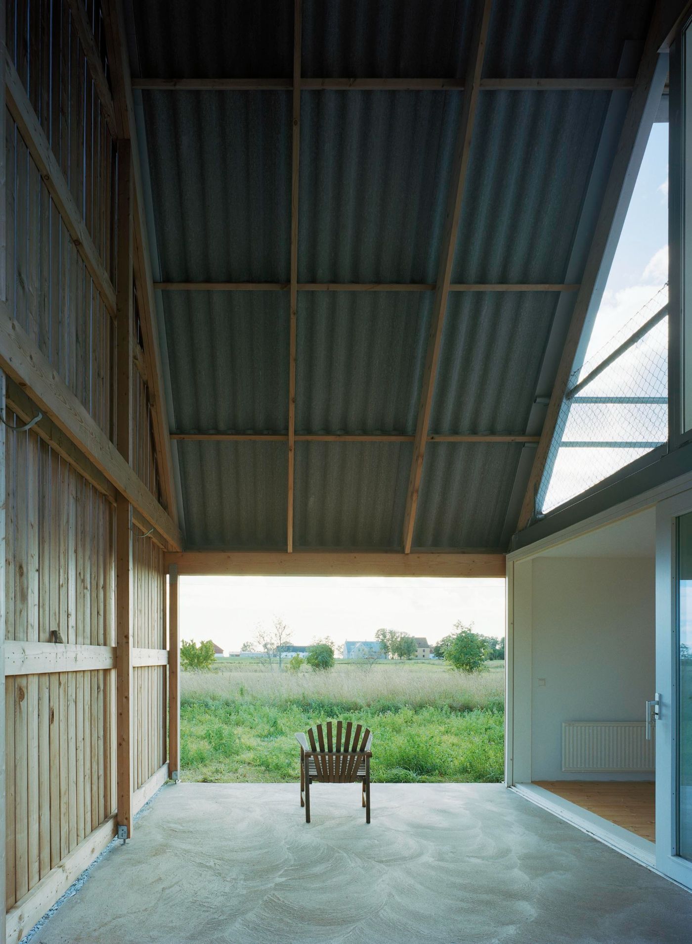 Sommarhus at Stora Gasmora by LLP Arkitektkontor — The entrance pavilion frames a single wooden Adirondack chair centered on a concrete floor, positioned as a threshold to the open meadow beyond.