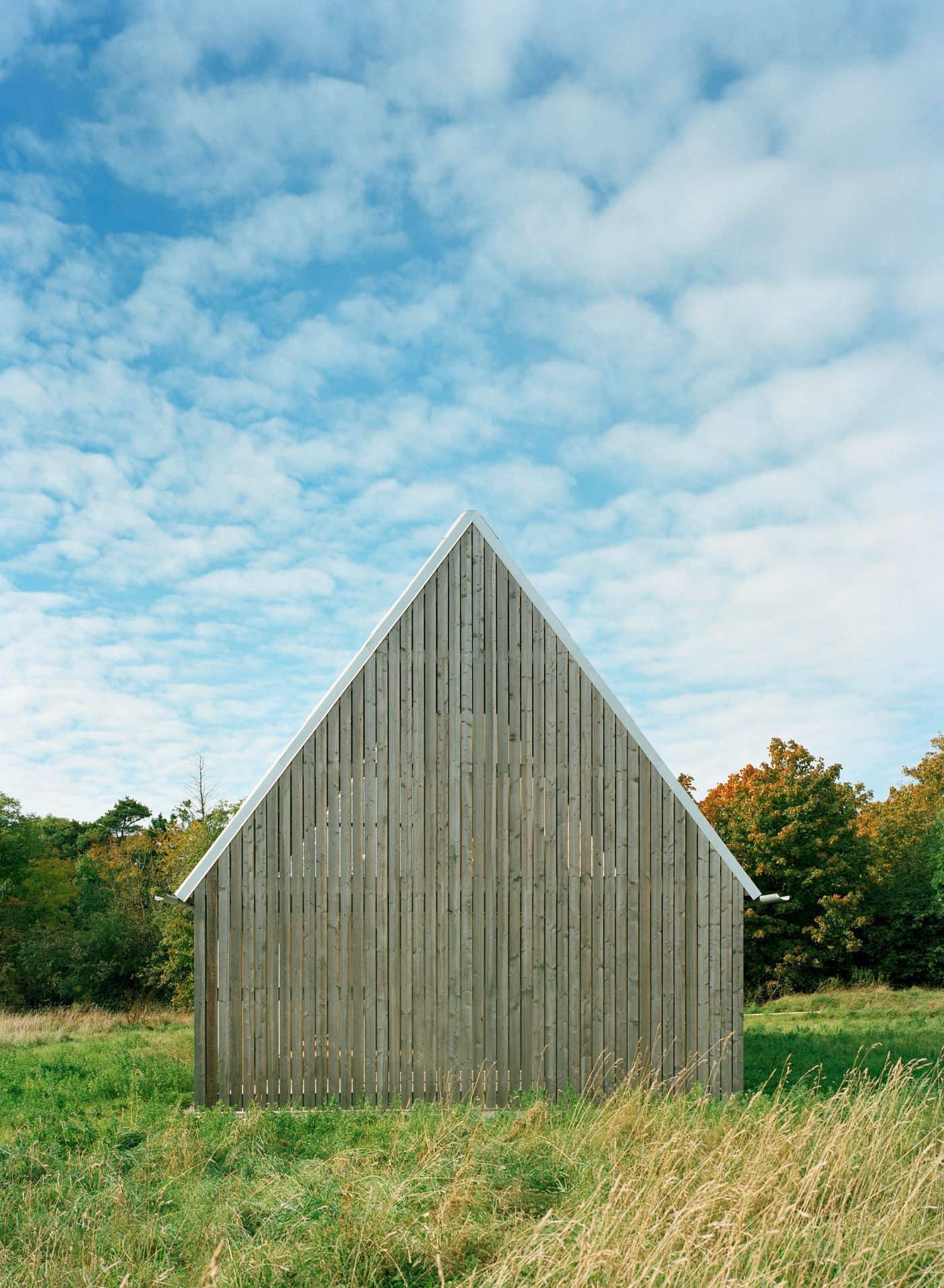 Sommarhus at Stora Gasmora by LLP Arkitektkontor — A pitched-roof structure clad in weathered larch timber dominates the meadow landscape, its gabled form rendered in vertical timber boards with...