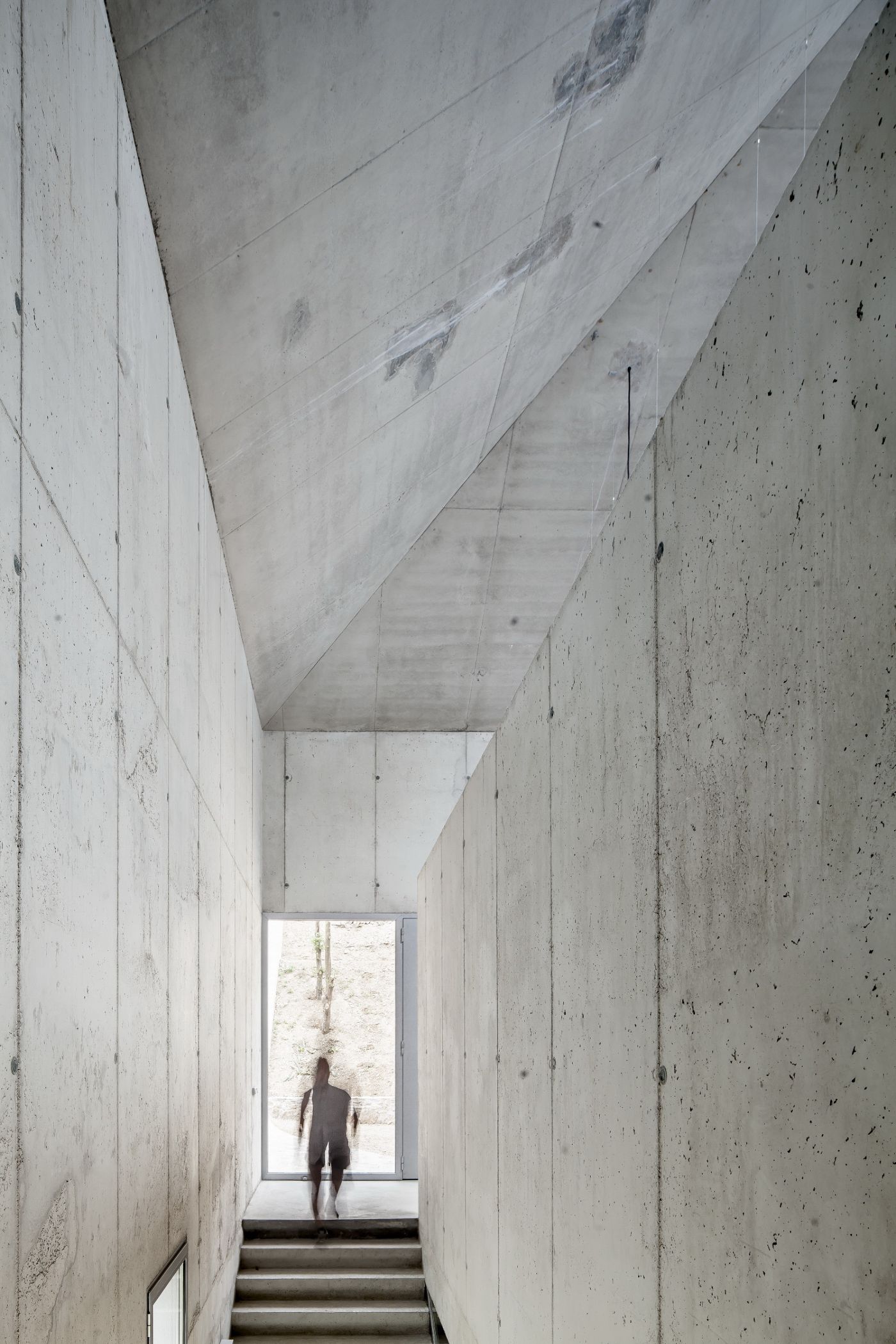 Arranz Bravo Studio by Garcés - De Seta - Bonet Arquitectes — A narrow concrete corridor descends toward a glazed threshold, framing a solitary figure silhouetted against daylight.
