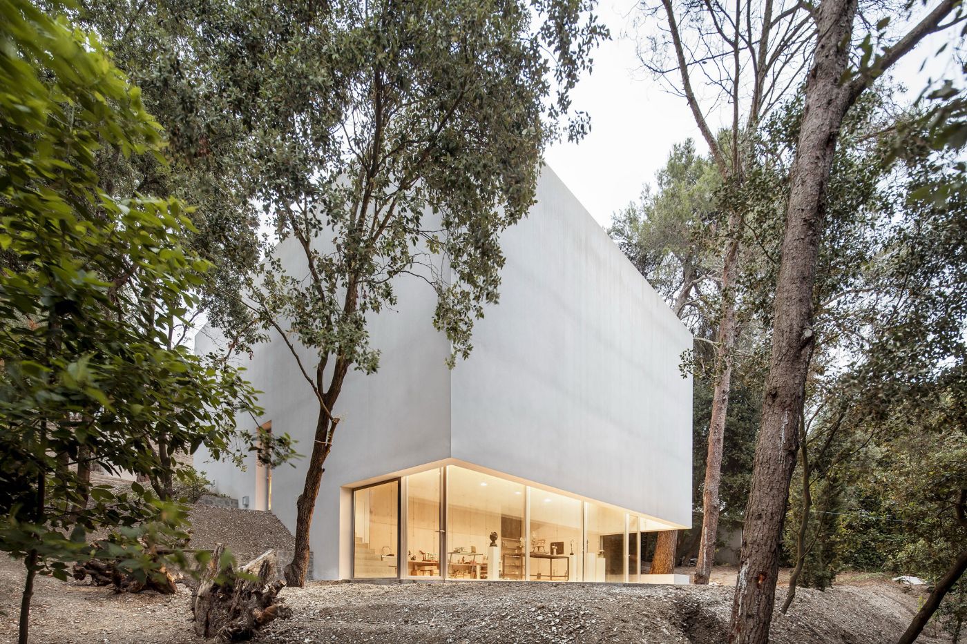 Arranz Bravo Studio by Garcés - De Seta - Bonet Arquitectes — A monolithic white rendered structure with a pitched roof rises among tall eucalyptus trees, its geometric form cutting a clean silhouette against the...