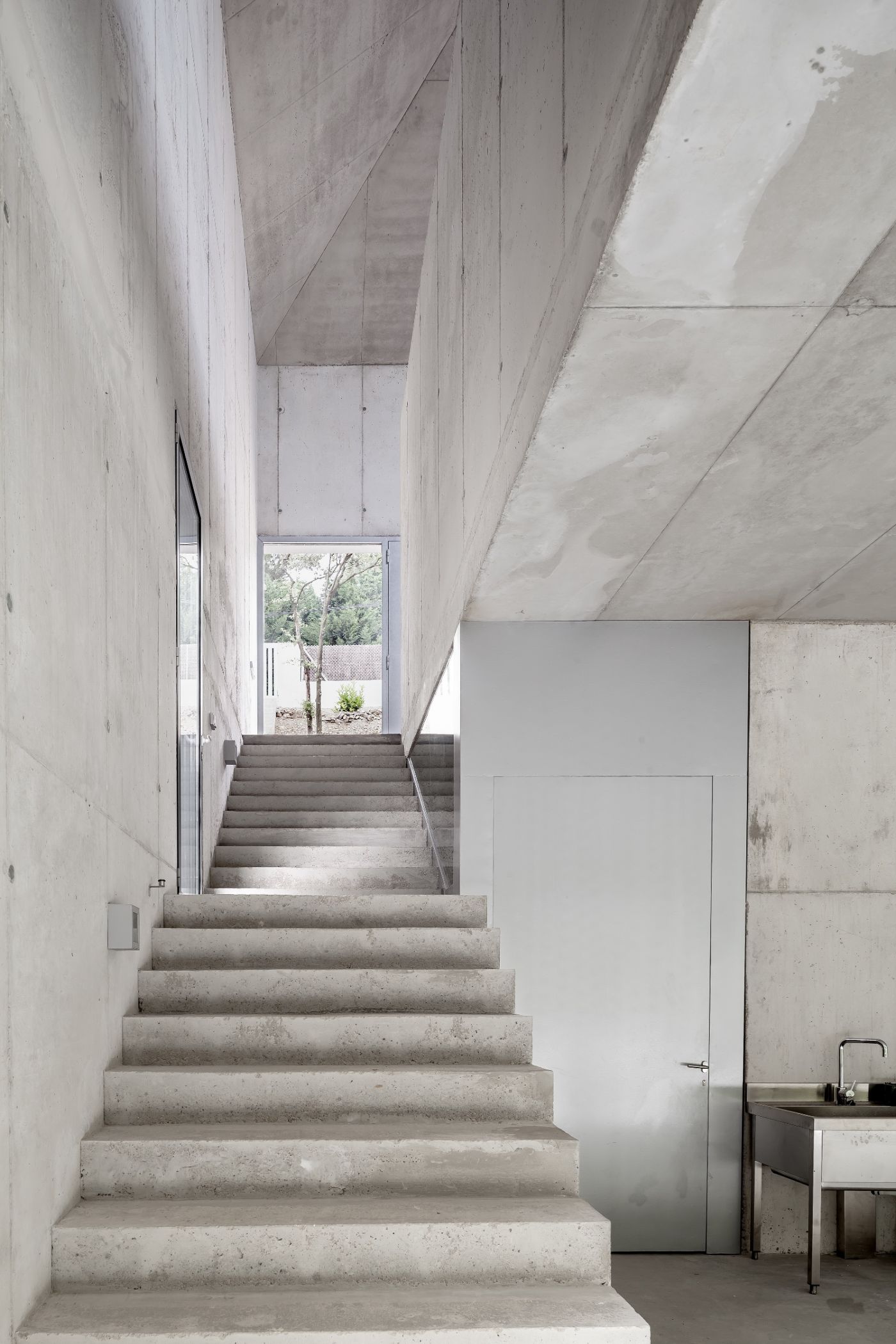 Arranz Bravo Studio by Garcés - De Seta - Bonet Arquitectes — A monolithic concrete staircase ascends through a narrow passage between board-formed concrete walls, leading toward a distant window framing trees and garden.