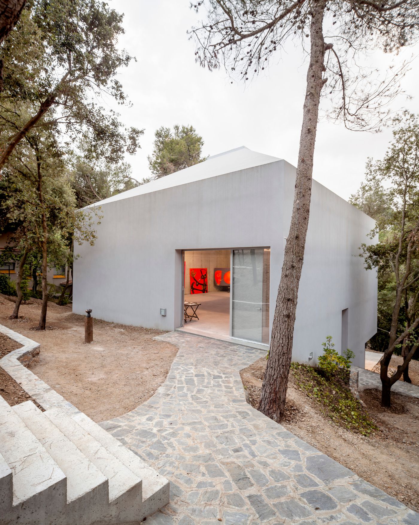 Arranz Bravo Studio by Garcés - De Seta - Bonet Arquitectes — A pitched-roof studio emerges from a pine forest clearing, its monolithic white rendered volume anchored by a continuous glazed threshold.