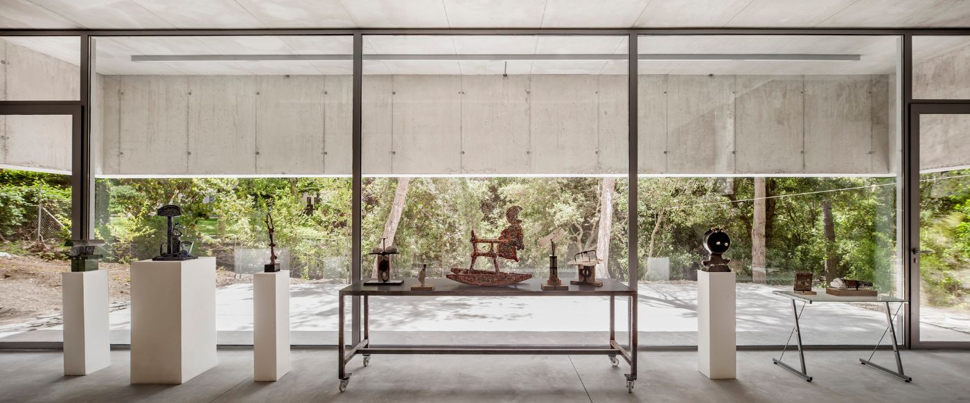 Arranz Bravo Studio by Garcés - De Seta - Bonet Arquitectes — A glazed gallery pavilion frames sculptural works displayed on white pedestals and a central steel table within the interior volume.