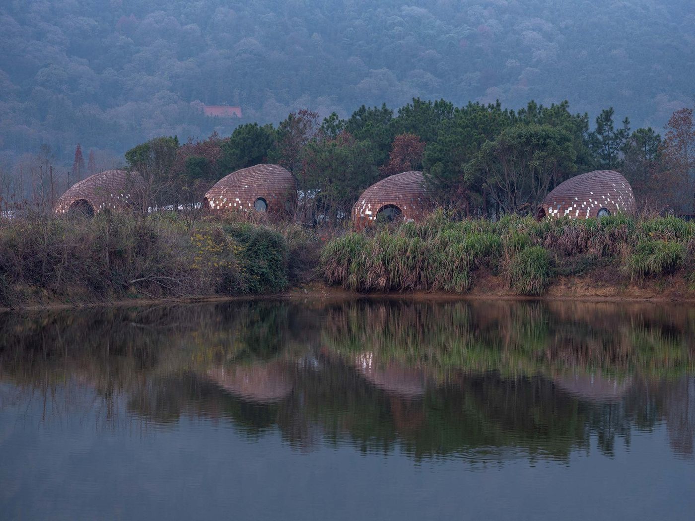 The Seeds Houses by ZJJZ Atelier — Three brick structures with curved ellipsoidal forms sit embedded in a densely forested landscape along a still waterway.