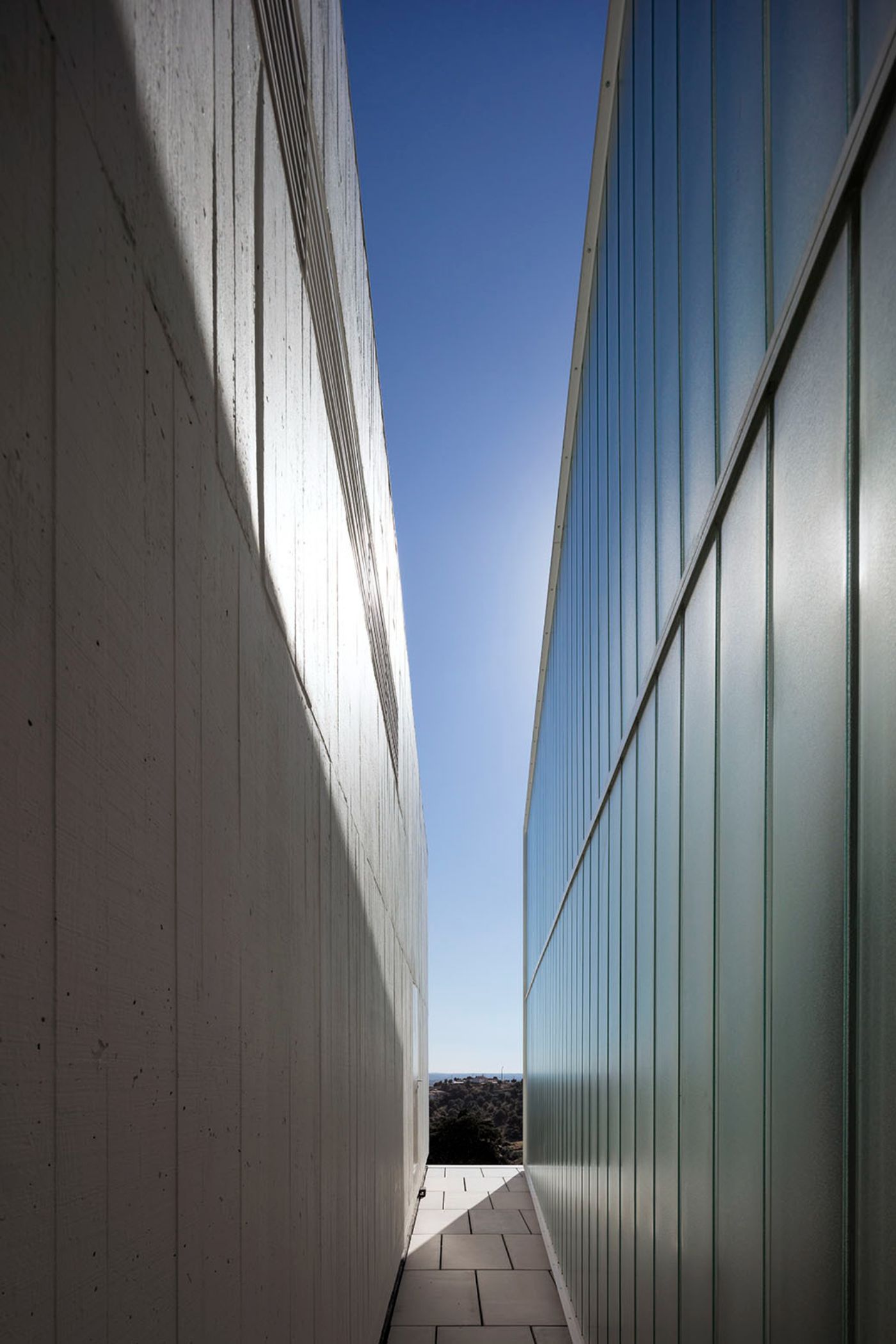 Abrantes Municipal Market by ARX Portugal — A narrow passage between two monolithic concrete walls converges toward a bright opening framed against clear blue sky.