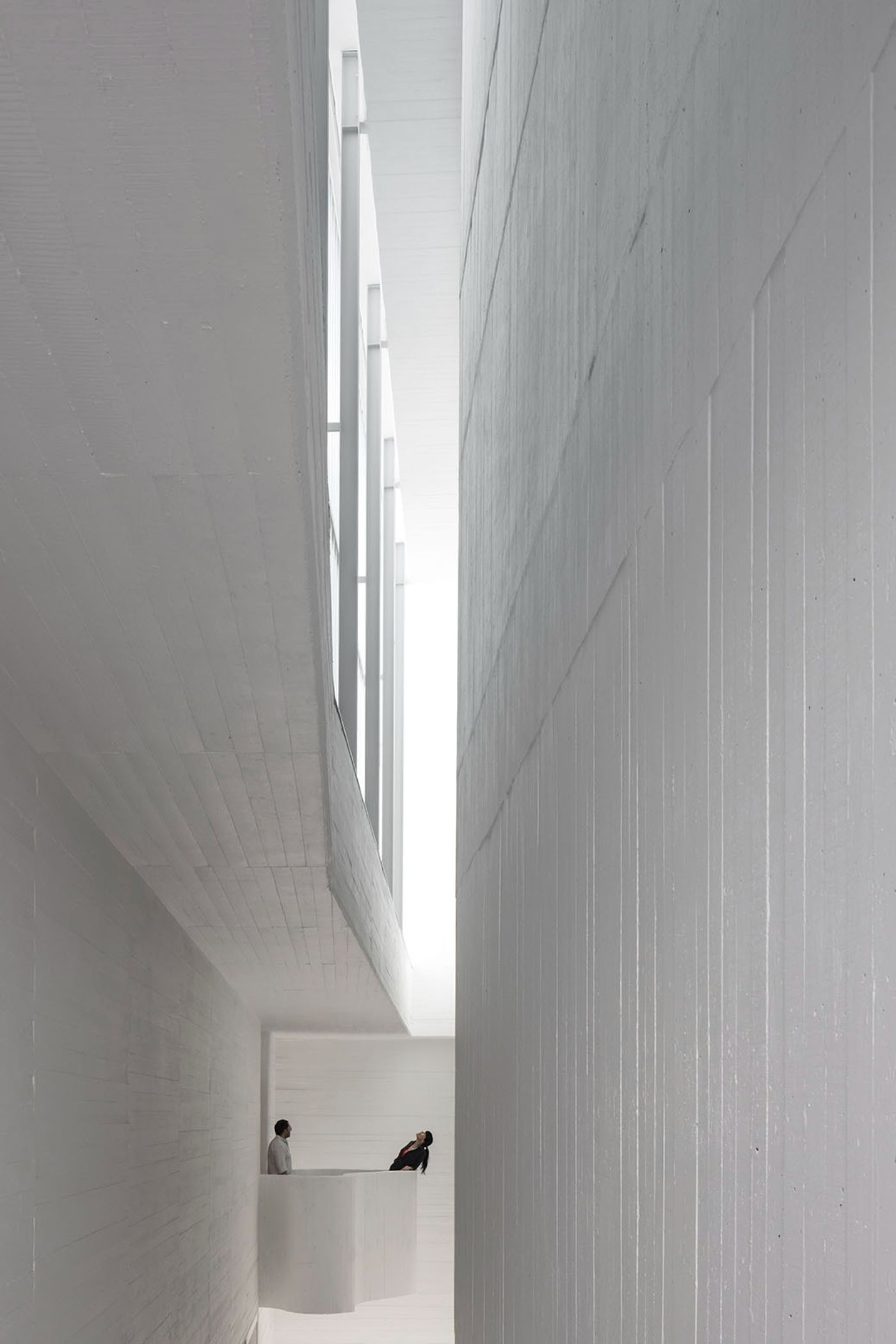 Abrantes Municipal Market by ARX Portugal — A narrow vertical passage between two monolithic concrete walls frames two figures in conversation at ground level.
